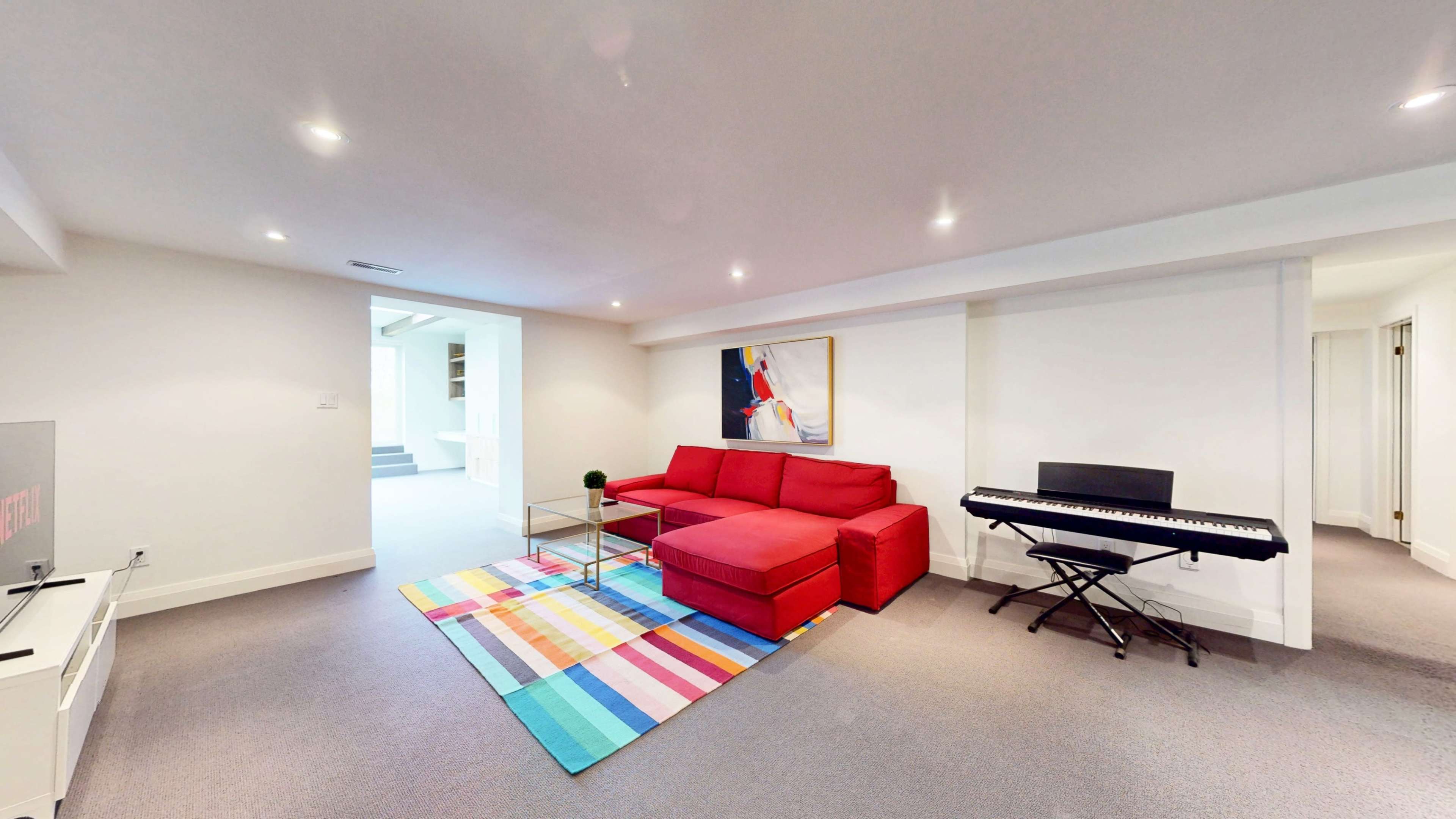 The image shows a well-lit basement living area featuring a red sectional couch, a colorful striped rug, a small glass coffee table, and a digital keyboard.