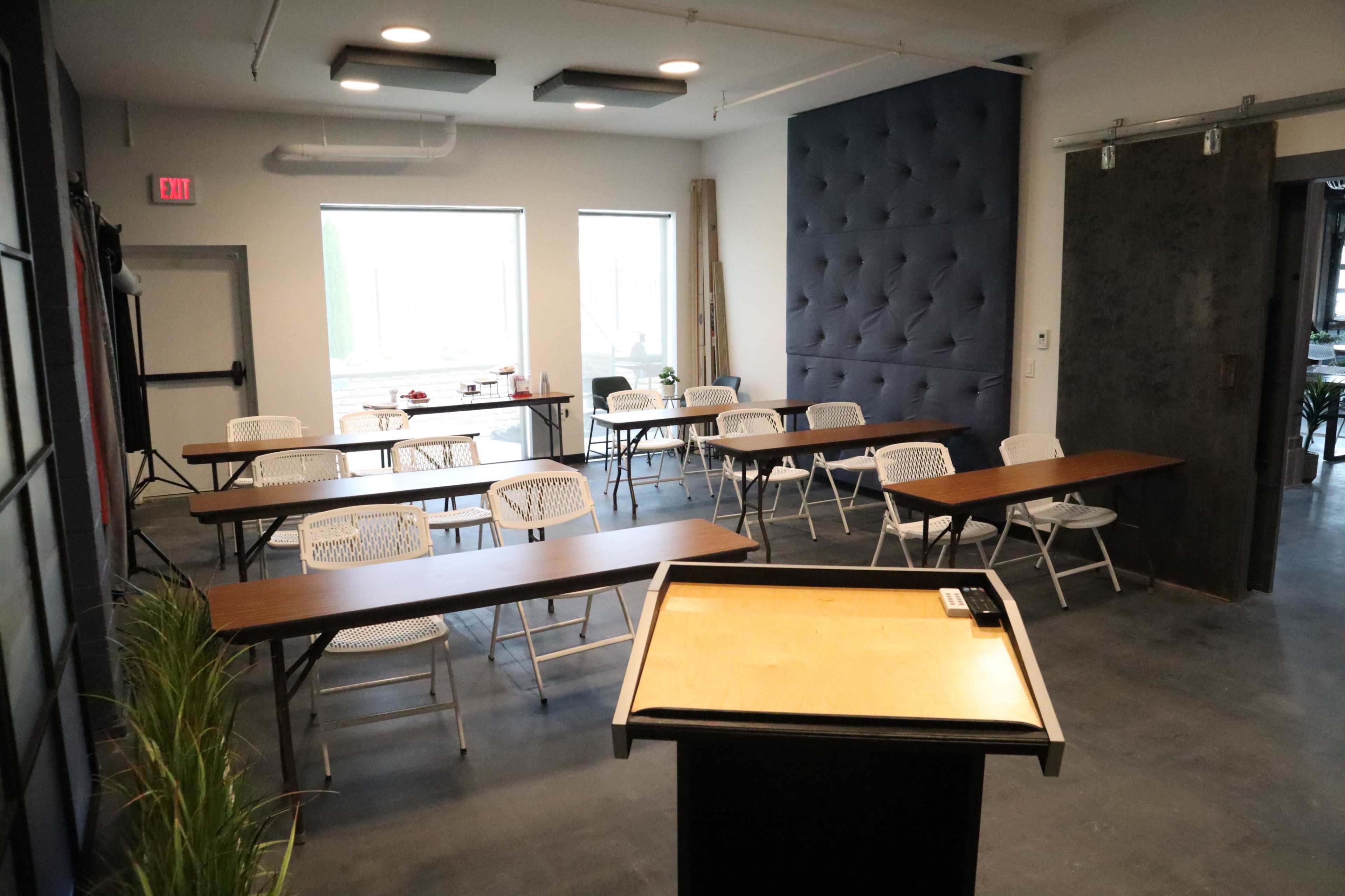 A classroom setup features several rows of tables and chairs, with a podium at the front and large windows providing natural light.