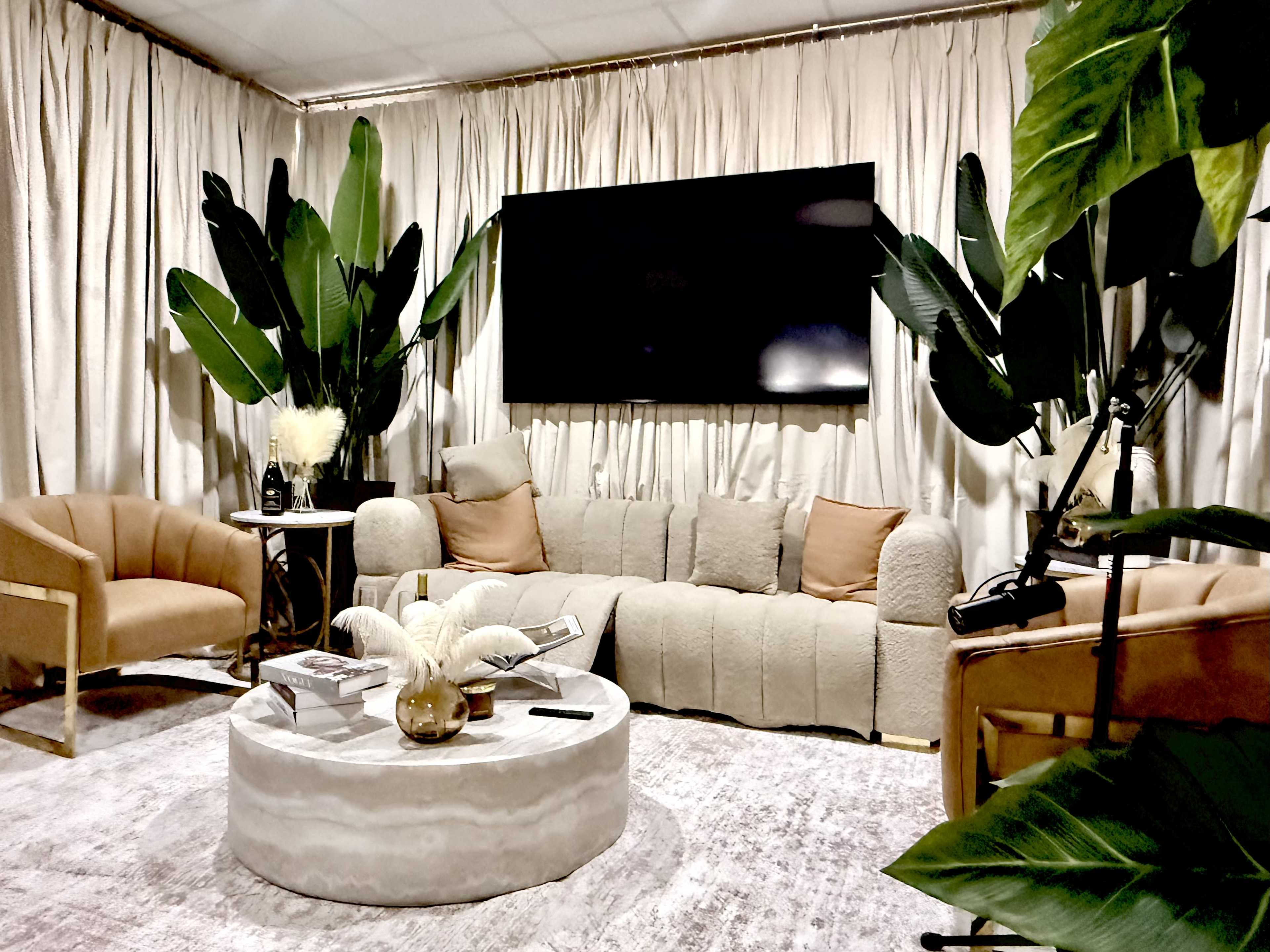 The image shows a cozy living room with a beige sofa, two armchairs, a round coffee table, and large green plants, all set against a backdrop of curtains and a wall-mounted TV.