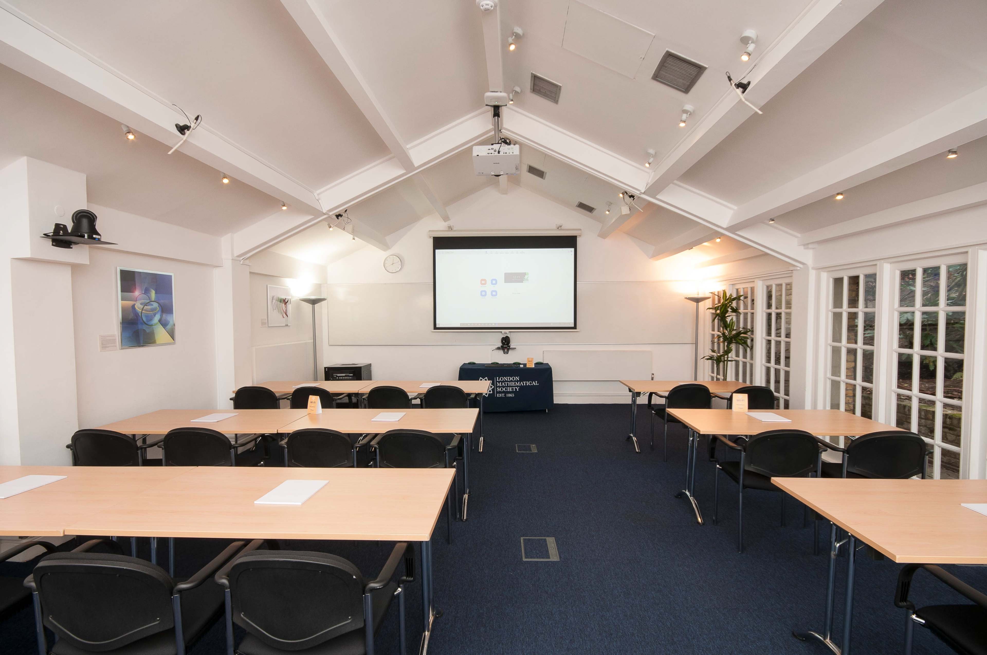 A meeting room features several tables arranged in rows facing a projection screen, with chairs neatly placed around the tables.