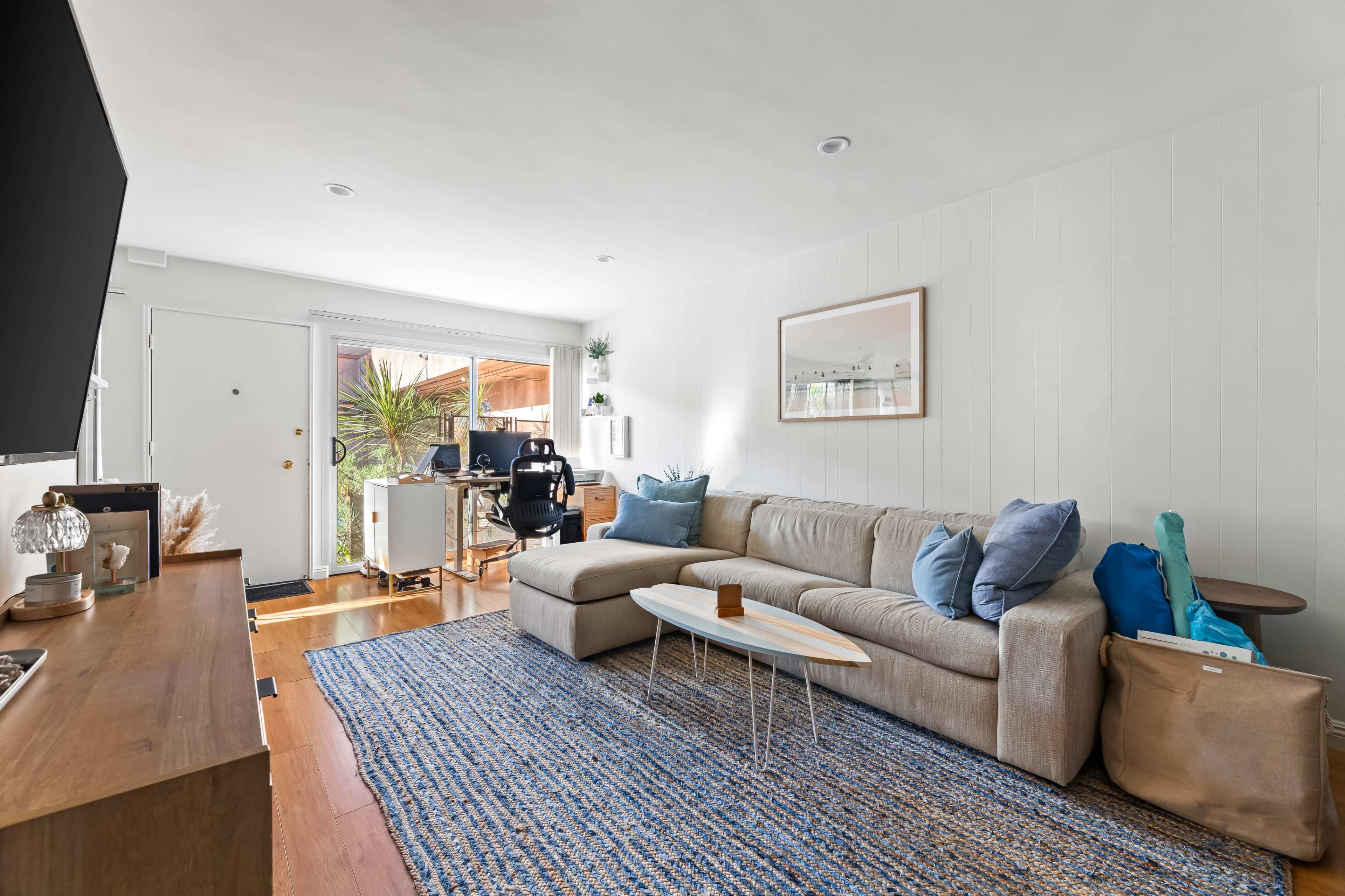 The living room features a beige sectional sofa, a wooden coffee table, a television on a console, and a patio door leading to an outdoor space.