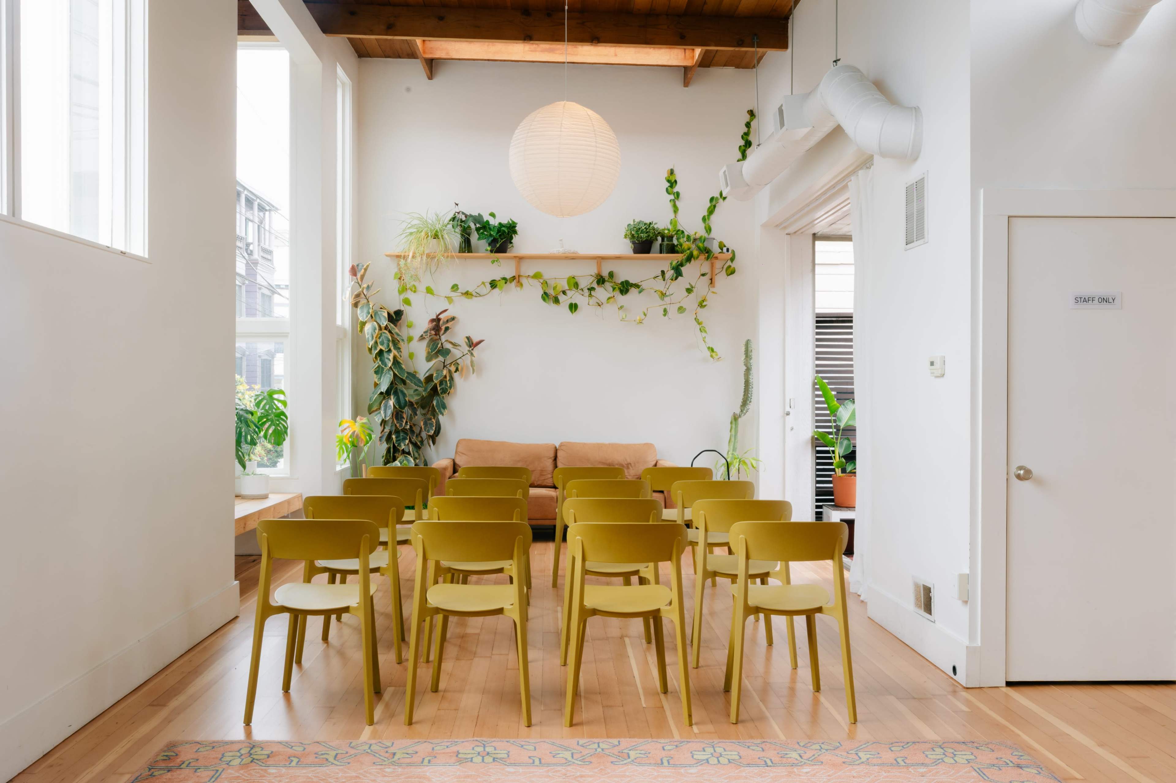 The image shows a modern indoor space with rows of yellow chairs, a brown couch, plants on shelves, and large windows allowing natural light.