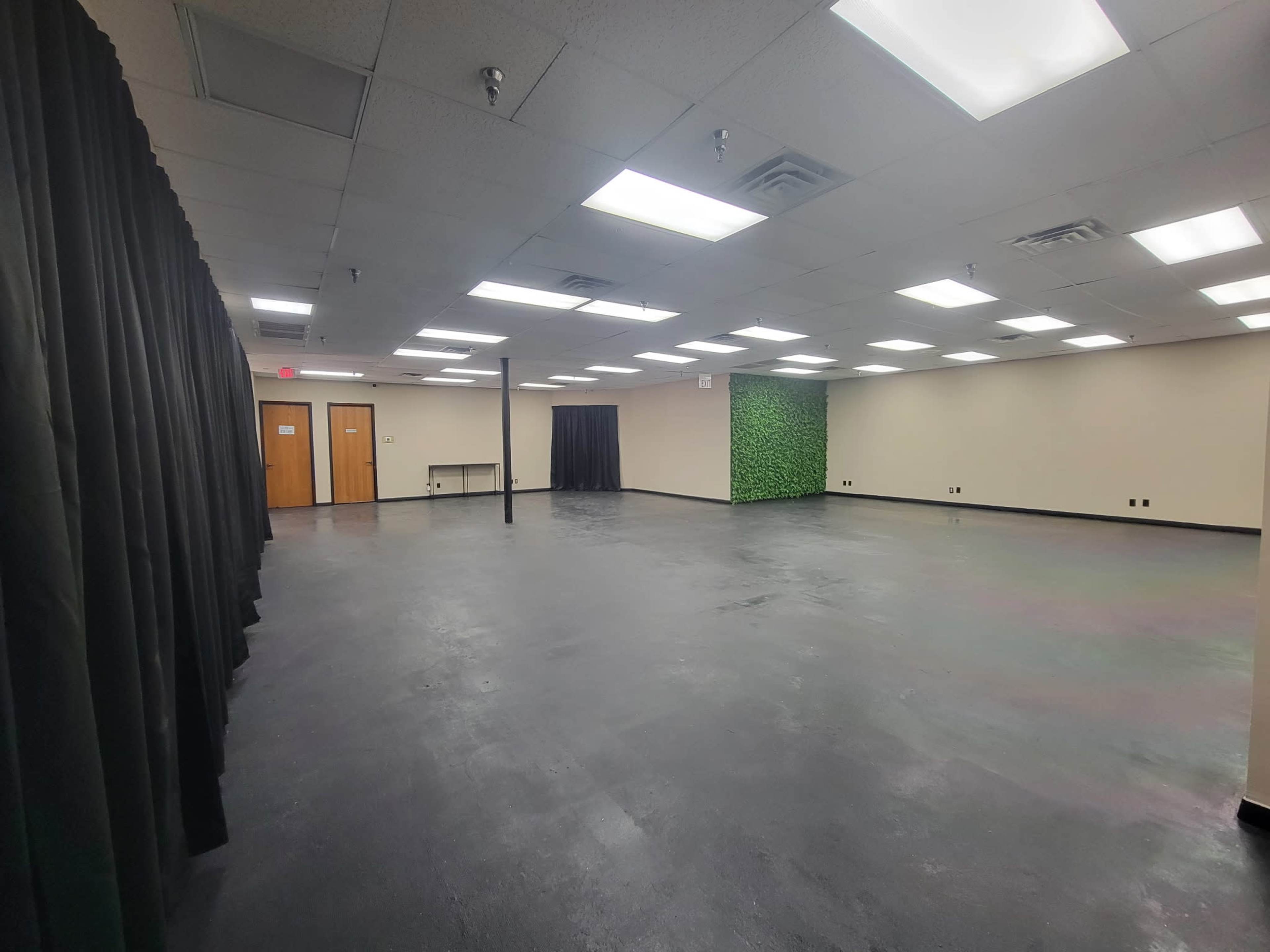 The image shows a spacious, empty room with a black curtain on one side, green foliage on another, and overhead fluorescent lights.