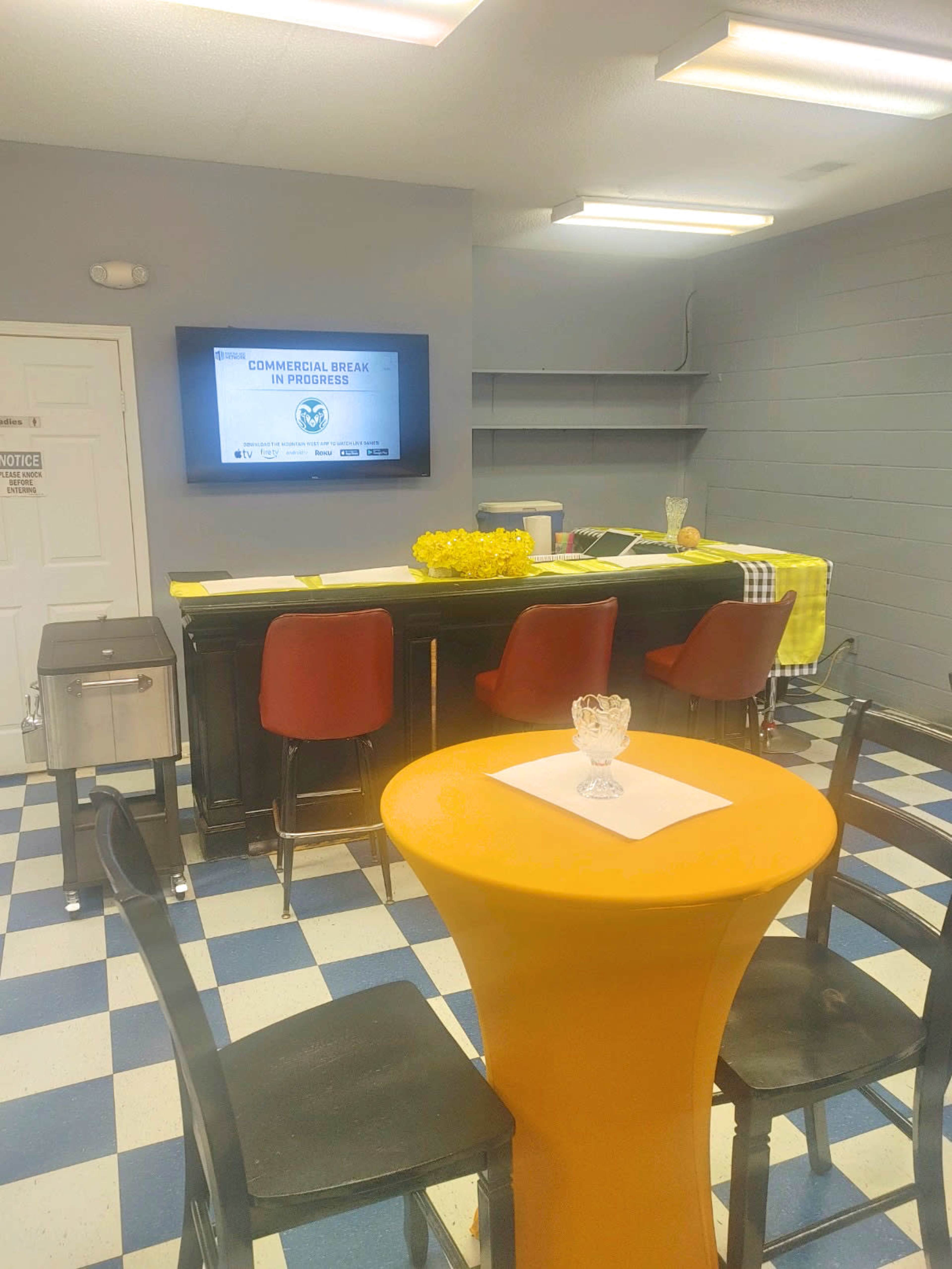 A small room with checkered floor tiles, featuring a bar counter with red chairs, a television displaying a commercial break sign, and a yellow tablecloth over a table.