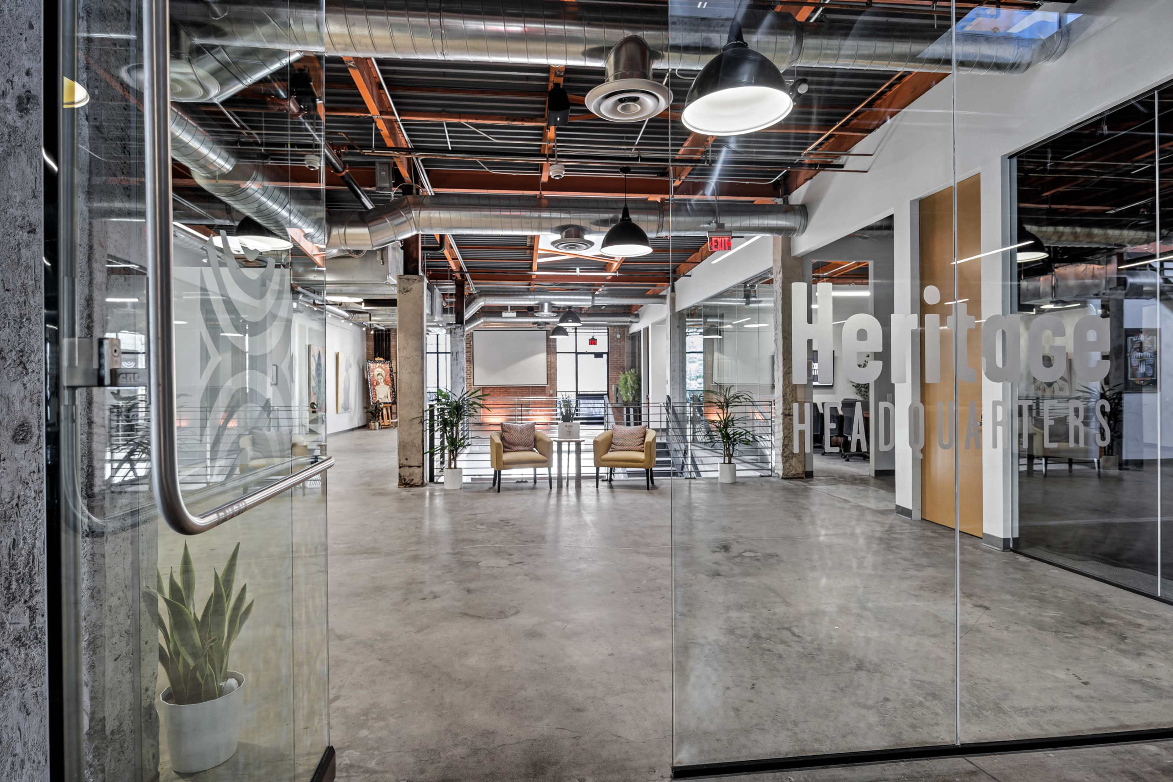 The image shows a modern office lobby with glass walls, concrete flooring, and contemporary seating arranged around plants.