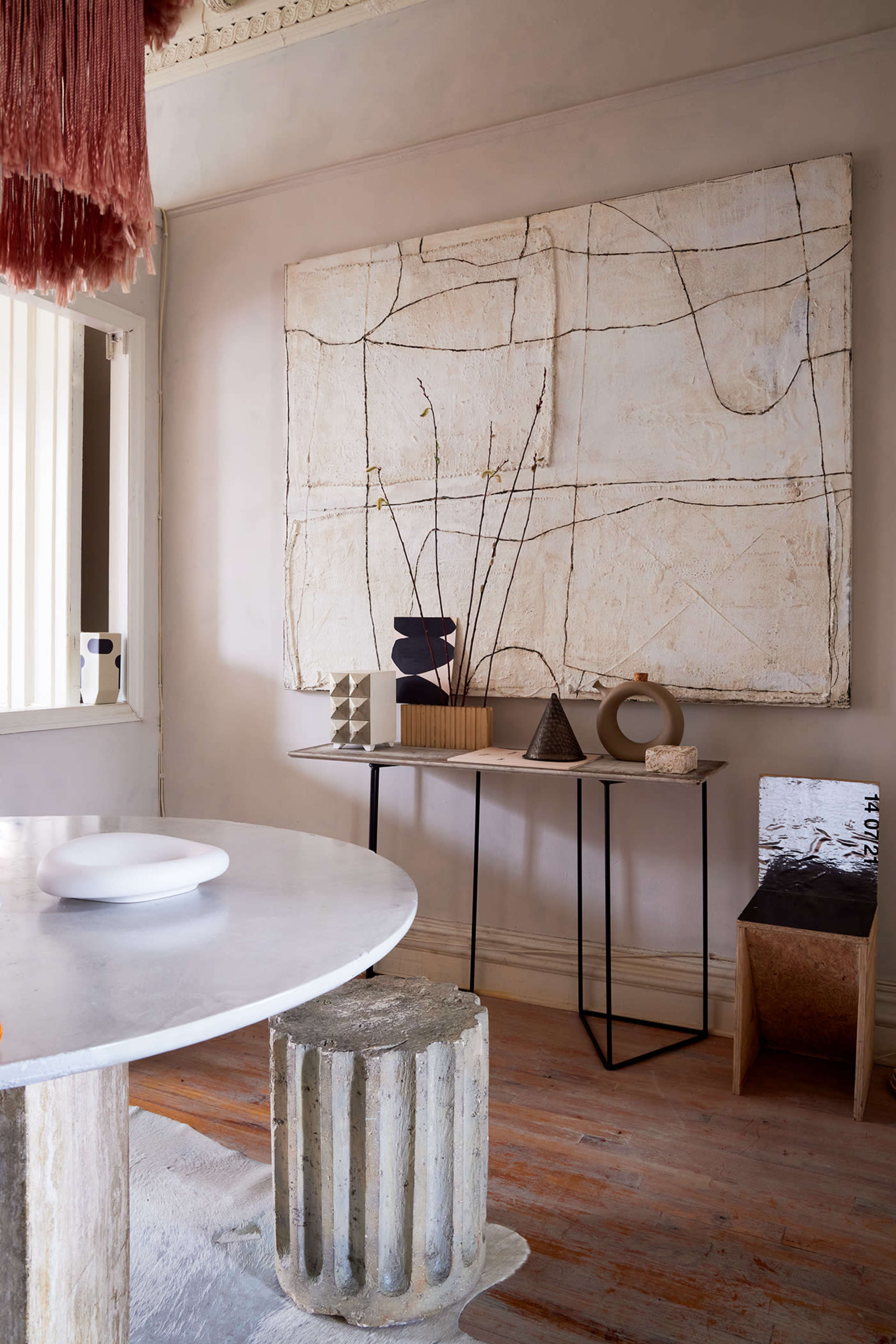 A marble table with a cylindrical base is positioned next to a wall featuring a large abstract artwork and a minimalist display of decorative objects on a black metal stand.