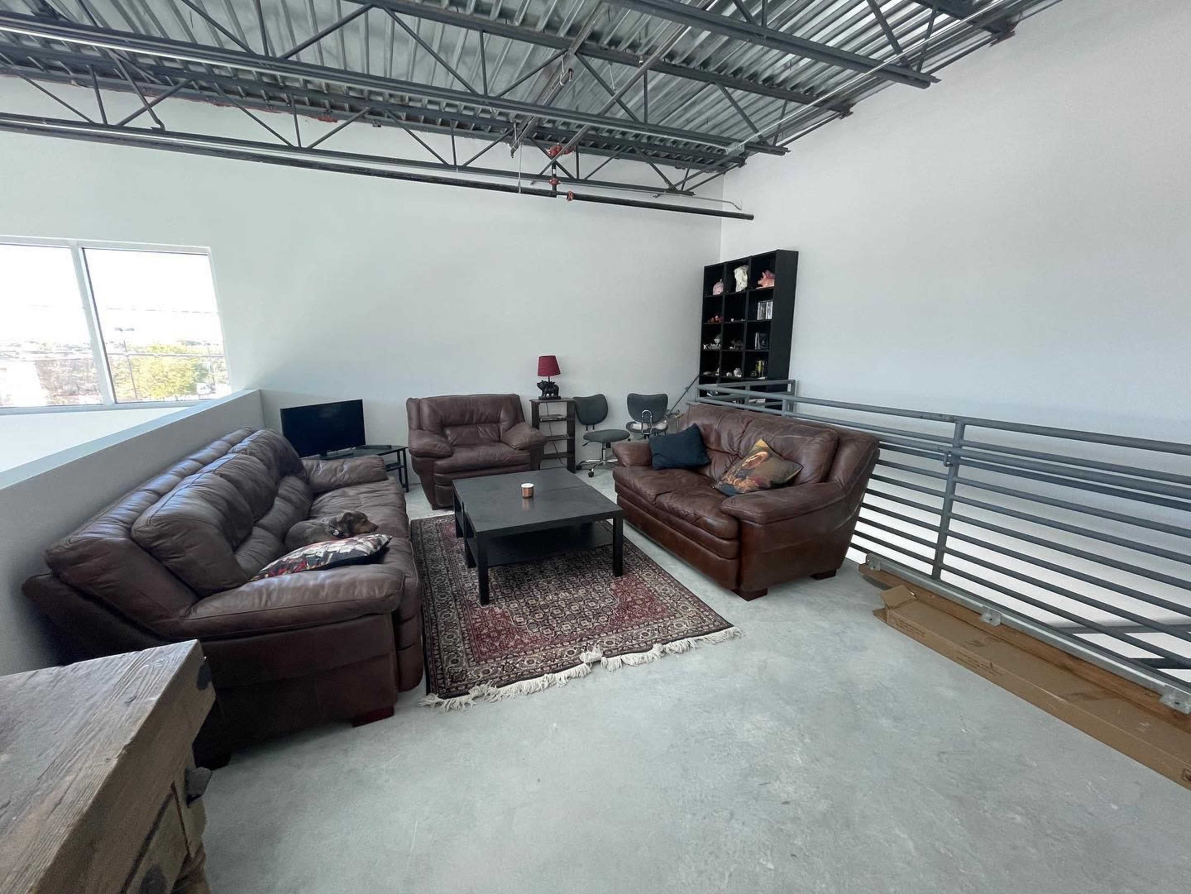 A spacious loft-style living area with two brown leather sofas, a coffee table on a patterned rug, and a bookcase in the corner.