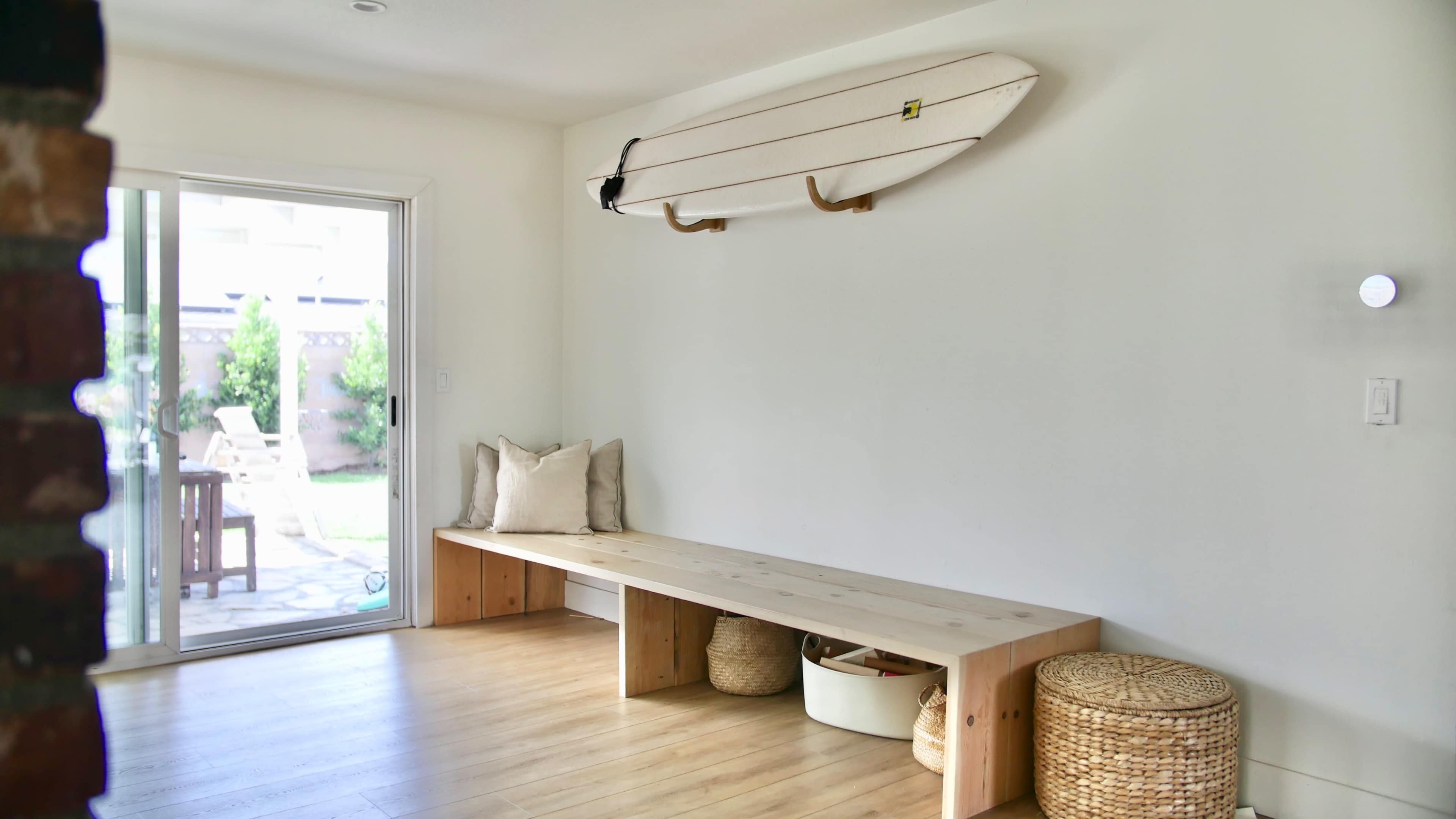 A minimalist room features a wooden bench, a surfboard mounted on the wall, and a sliding glass door leading to a garden.