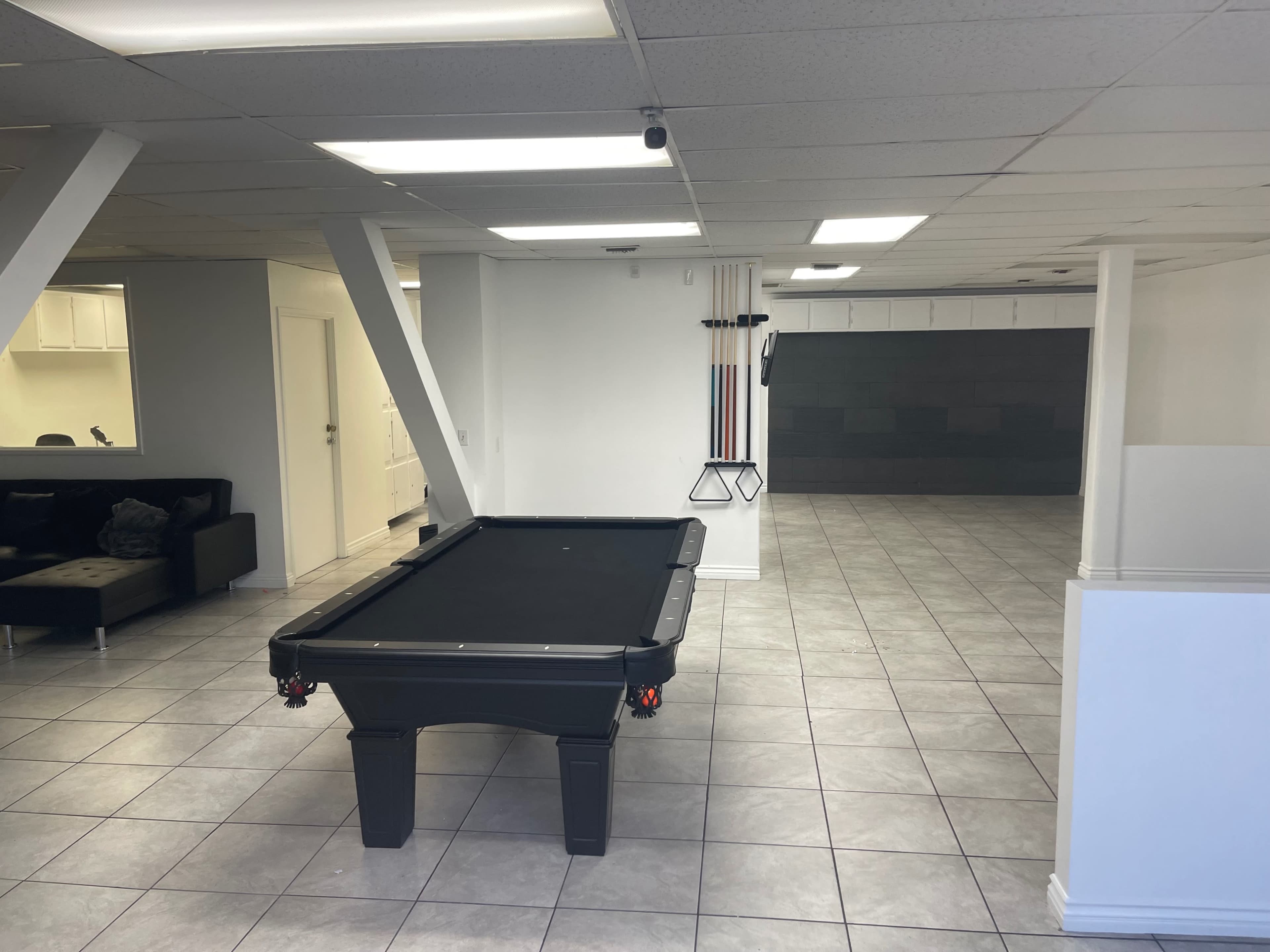 A spacious room featuring a black pool table in the center, with tiled flooring and minimal furnishings.