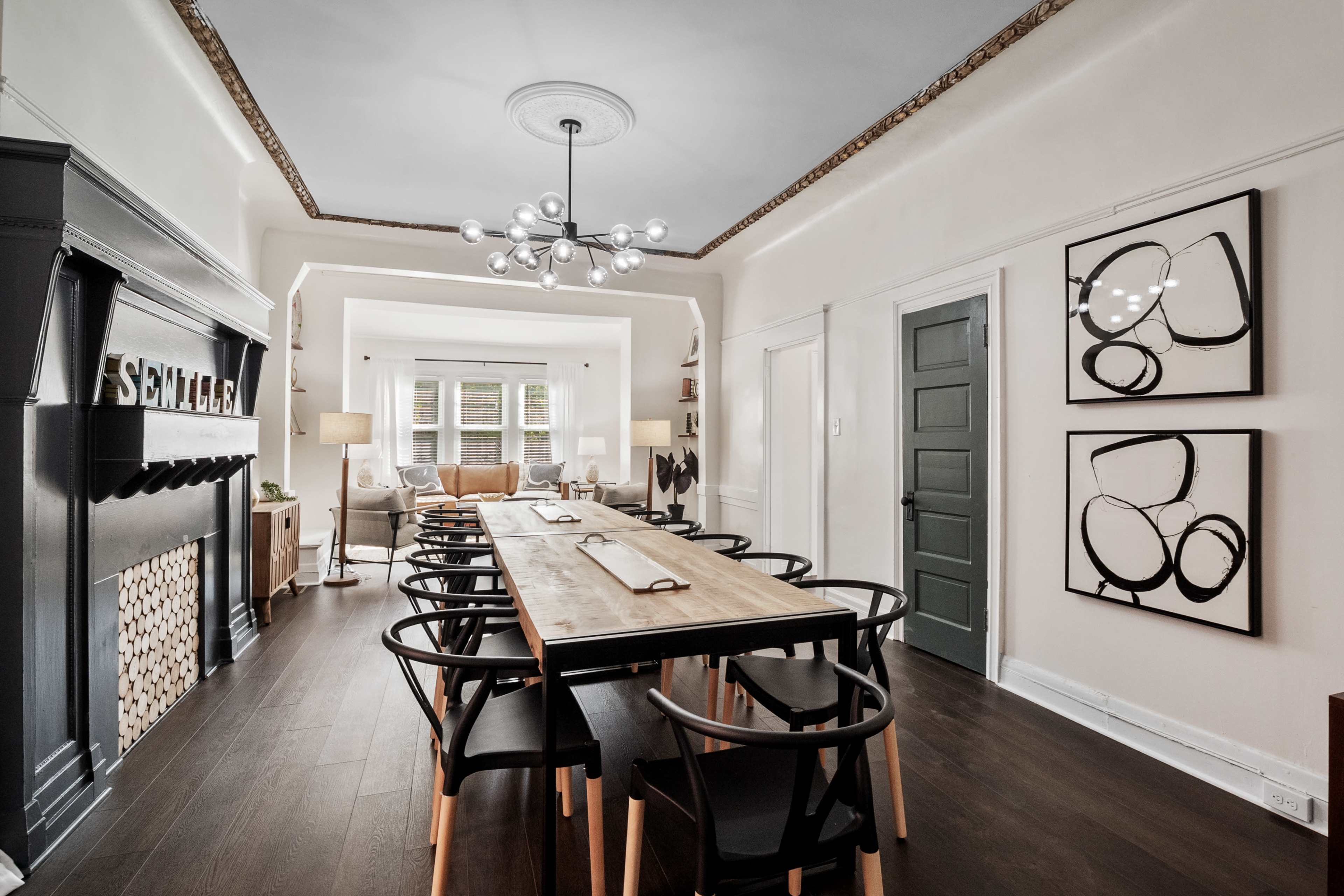 A long wooden dining table with black chairs is set in a well-lit room featuring a decorative fireplace and framed artworks on the walls.