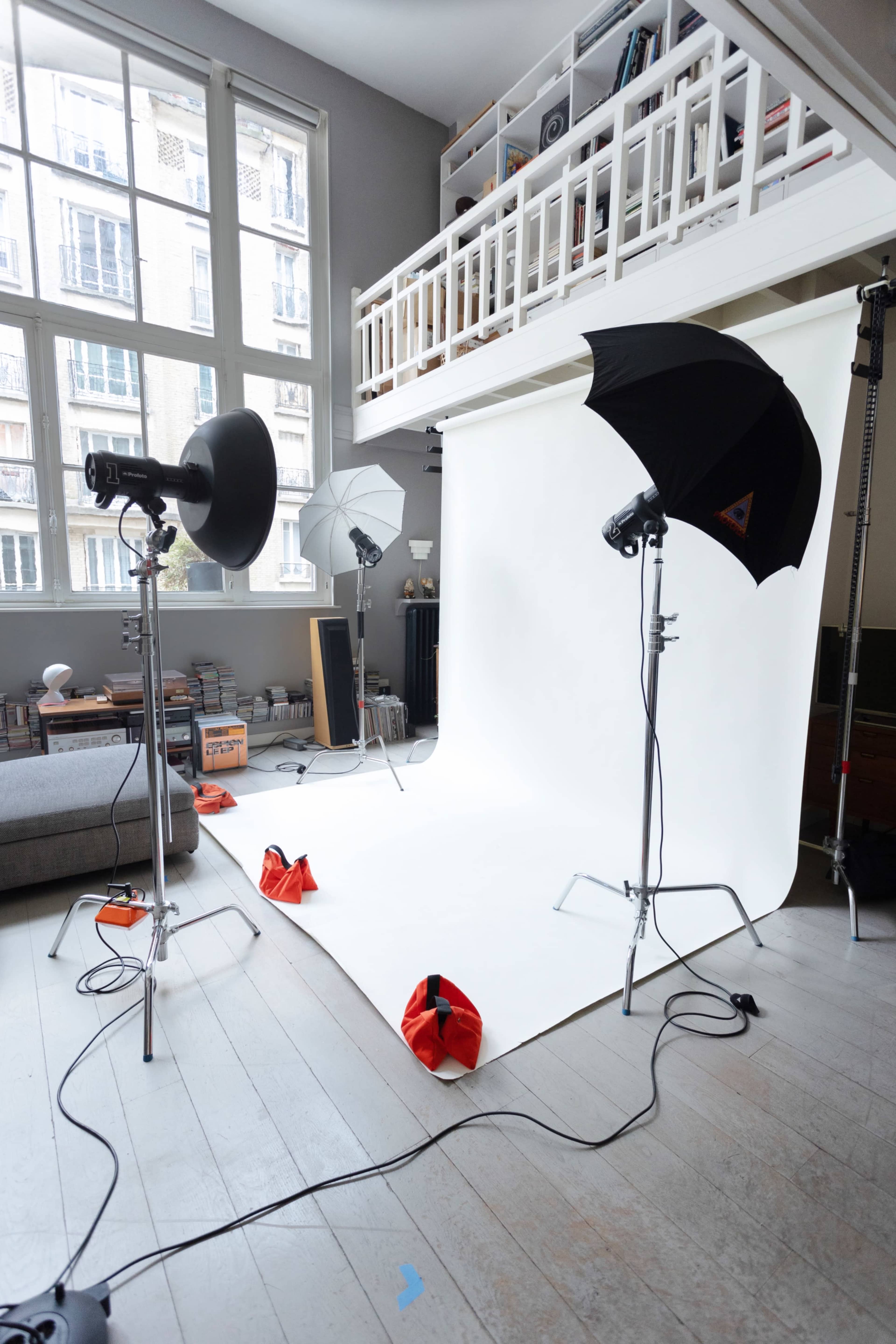 The image shows a photography studio setup in a room with large windows, featuring a white backdrop and several lighting equipment stands.