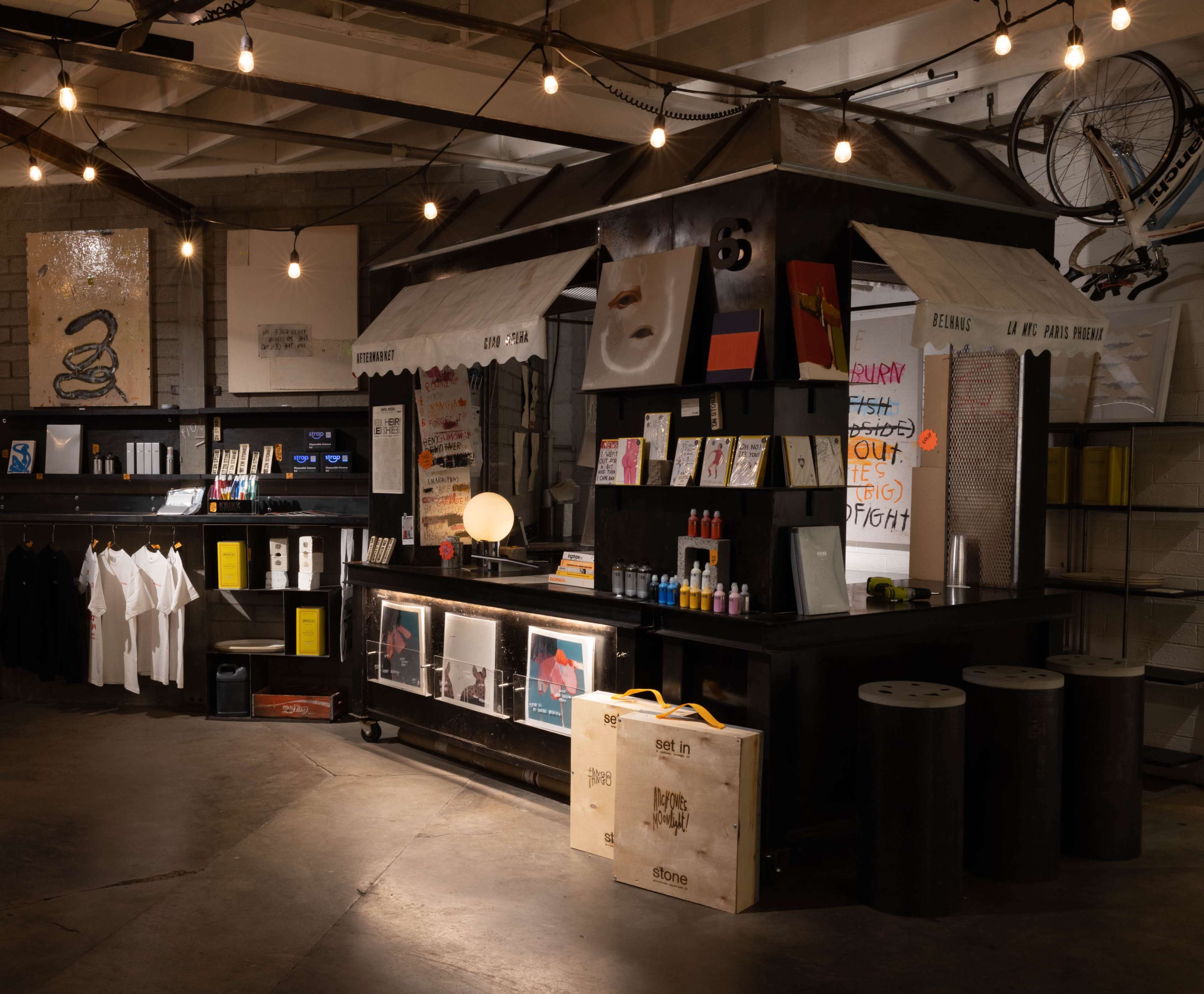 The image shows a stylish retail space with a dark wooden counter displaying various products, shelves filled with items, and dim string lights overhead.