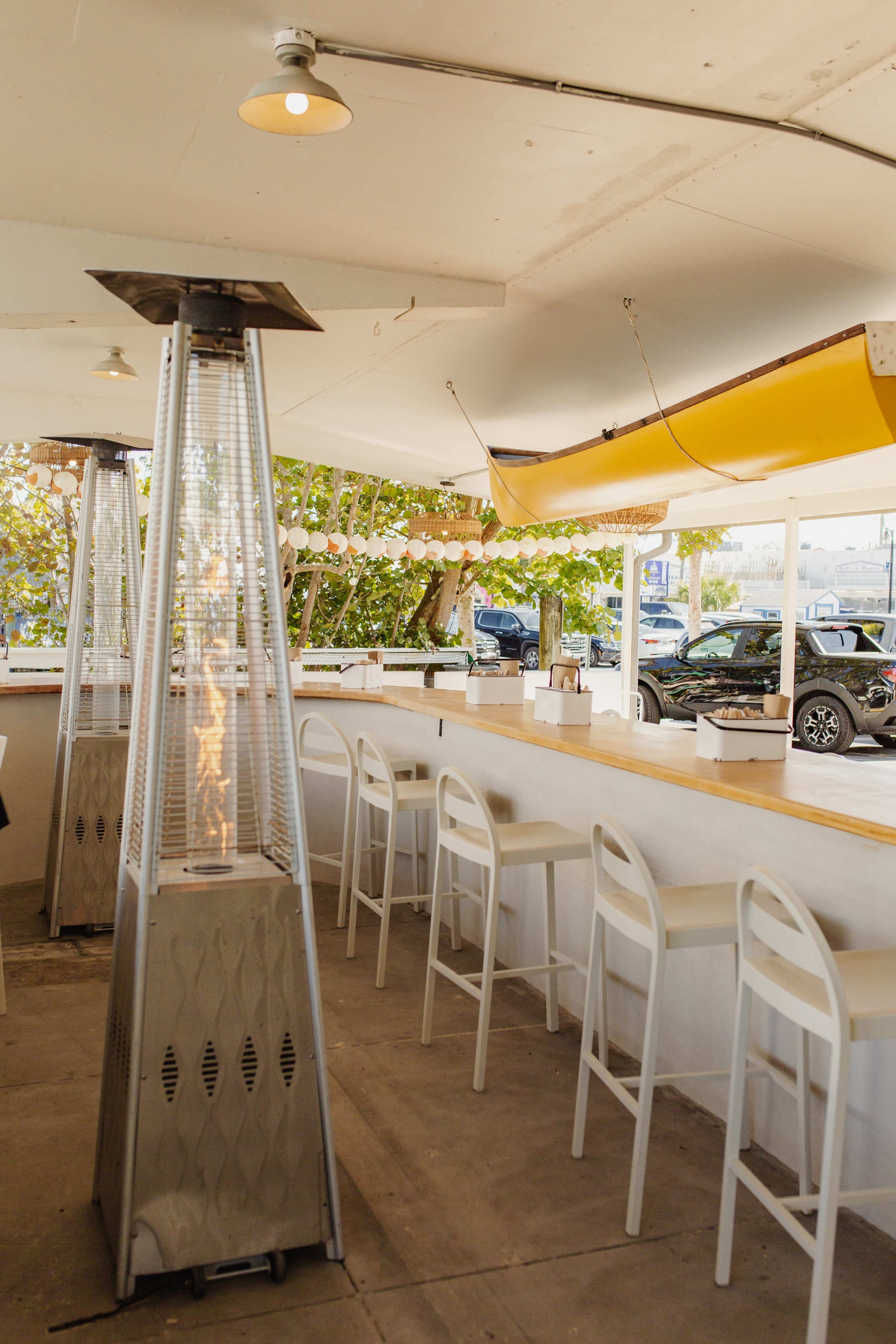 The image shows an indoor dining area featuring tall glass patio heaters and a long bar with white stools, illuminated by string lights and a kayak hanging overhead.