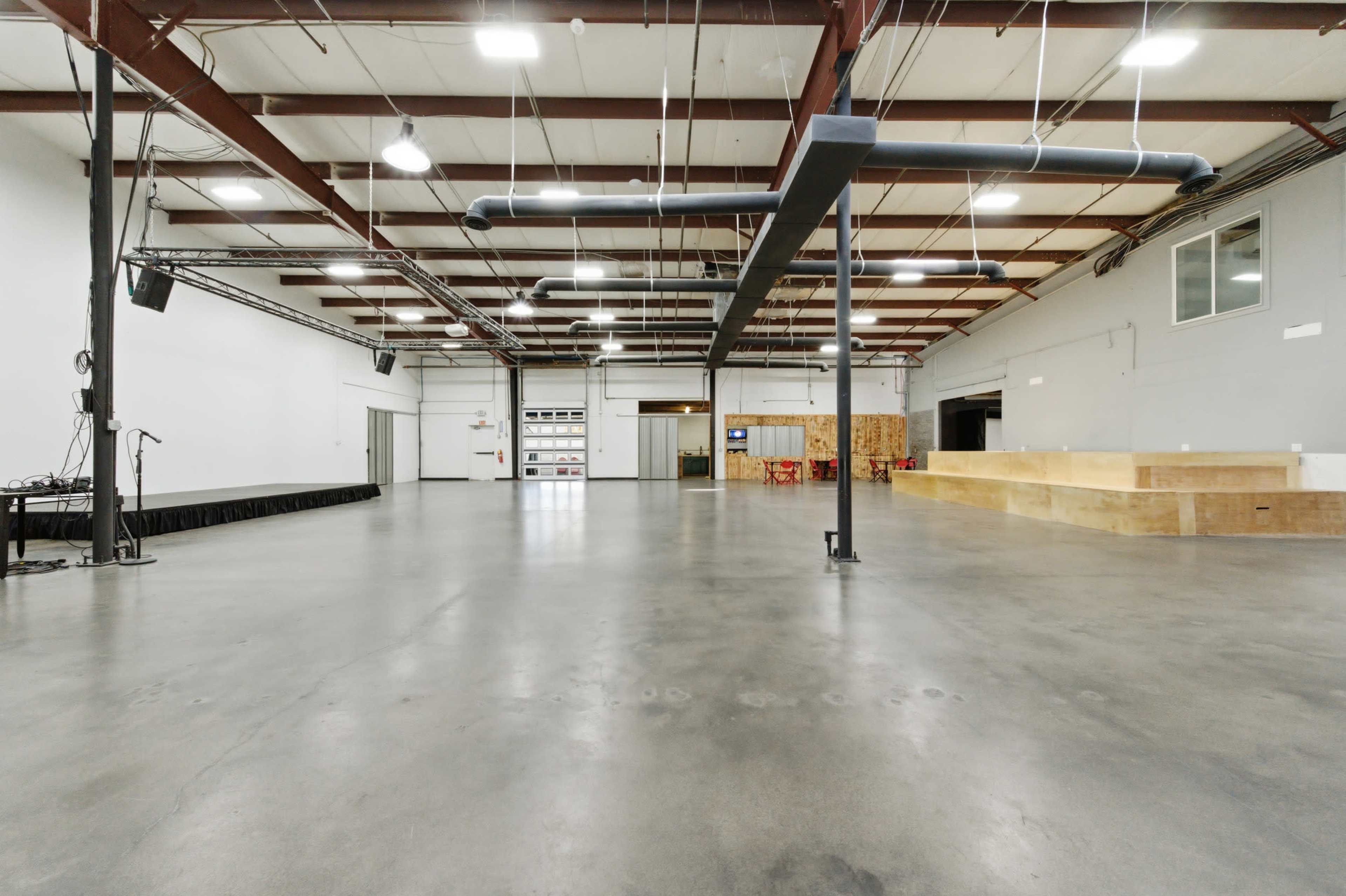 The image shows a spacious, industrial-style indoor venue with high ceilings, concrete flooring, and minimal furnishings.