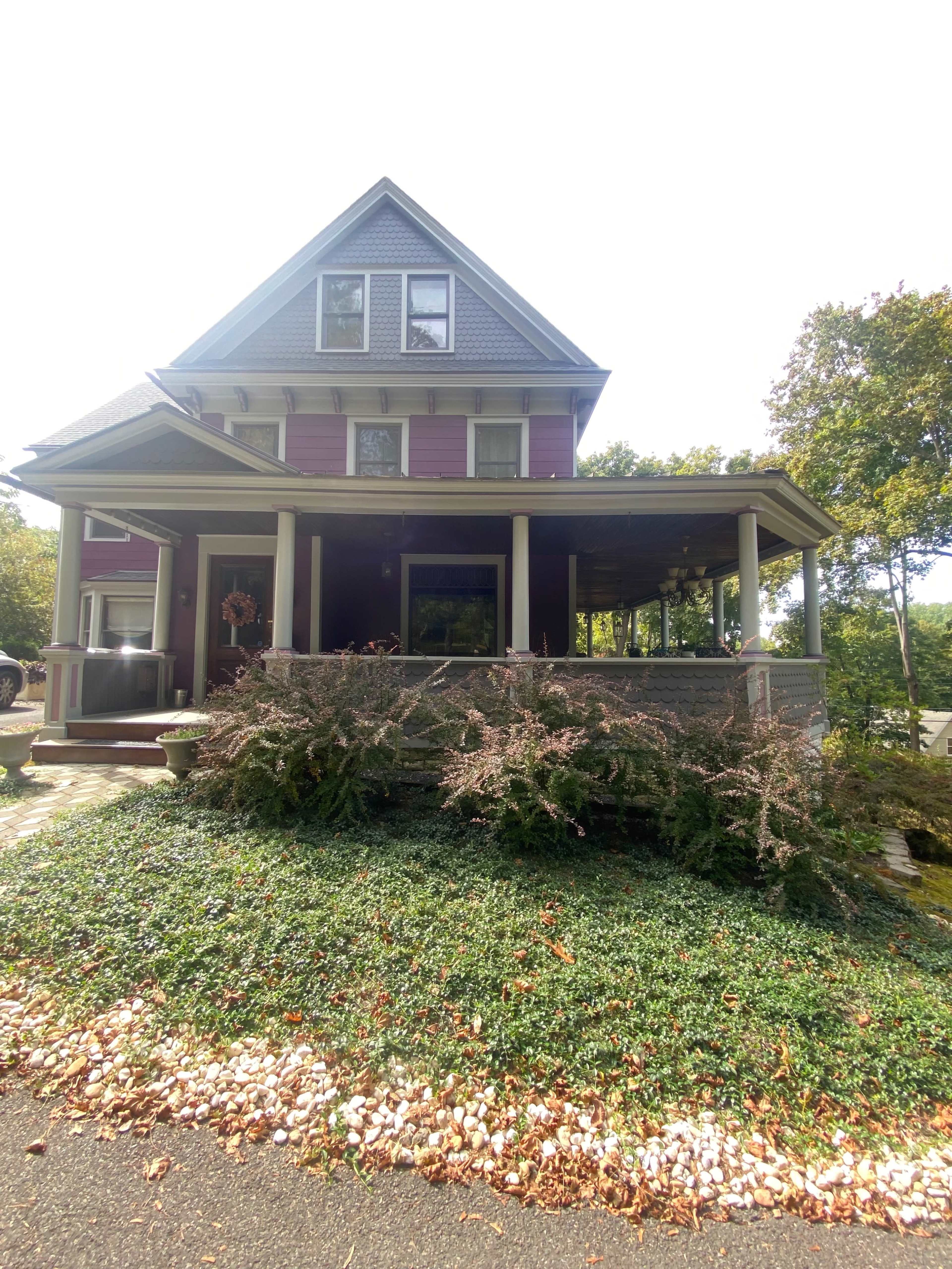 A two-story house with a gabled roof, featuring a porch and surrounded by green foliage and decorative stones.