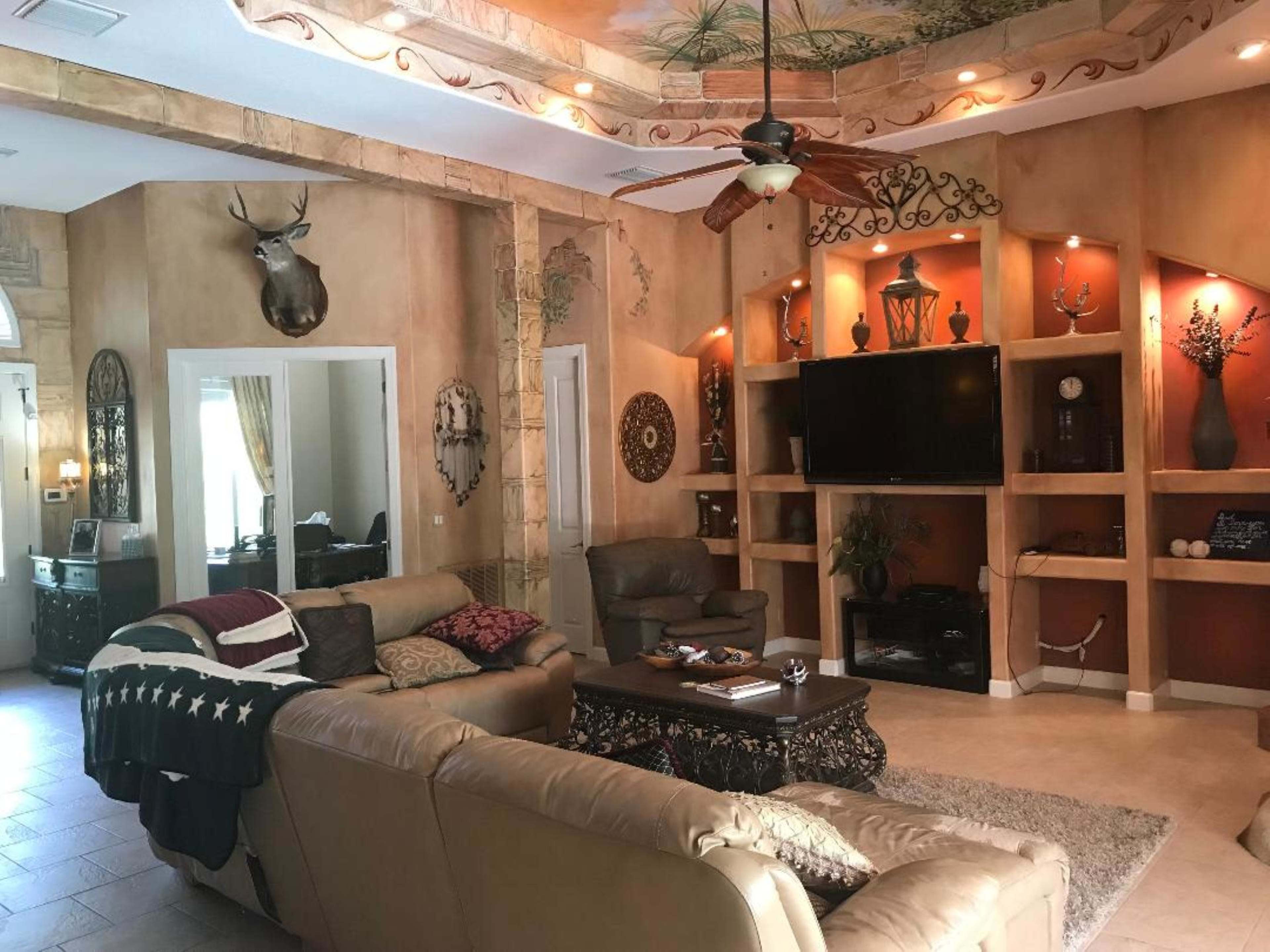 A living room features a large sectional sofa, a television mounted on a wall unit, and decorative elements including a mounted deer head and various wall accents.
