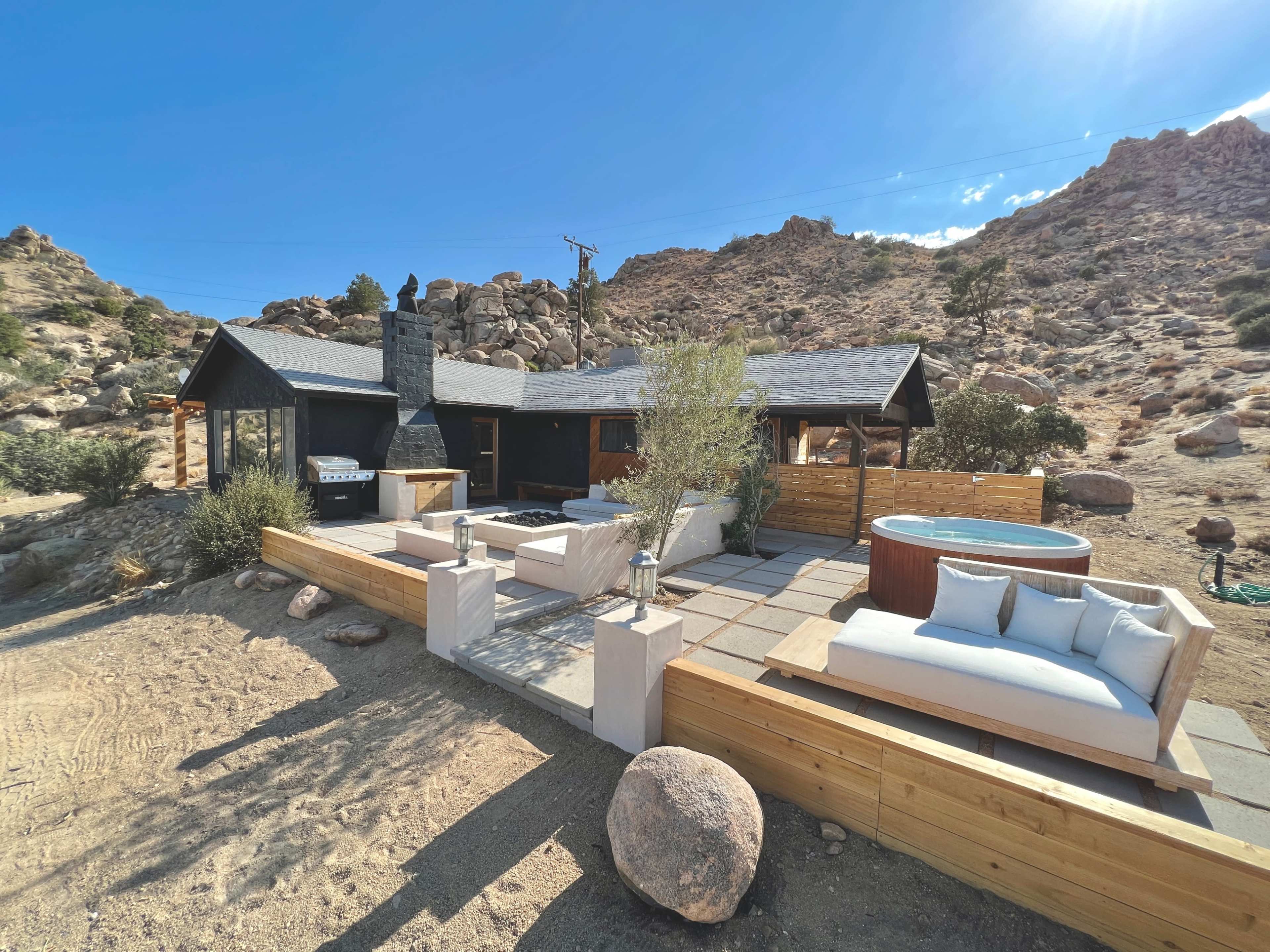 The image shows a modern house set against a rocky hillside, featuring a patio with seating, a hot tub, and a BBQ area.