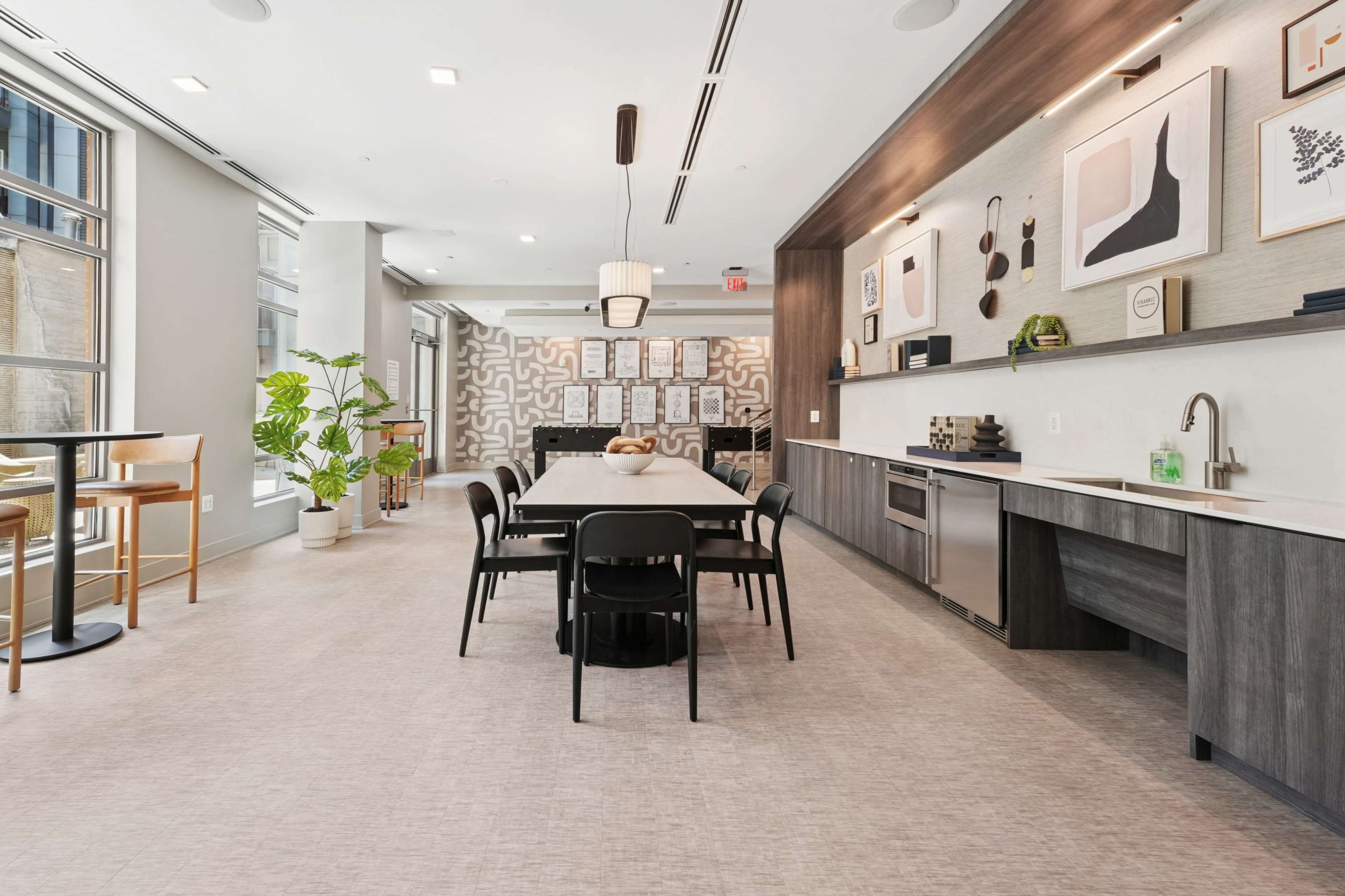 The image shows a modern kitchen and dining area with a long table, black chairs, and a counter with kitchen appliances and decorative artwork on the walls.