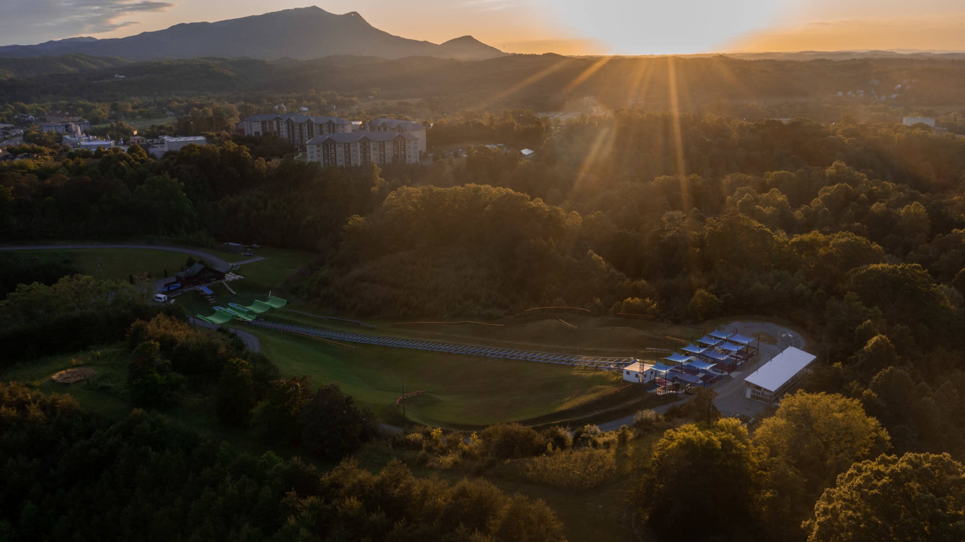 A scenic landscape at sunset, featuring rolling hills, a facility nestled among trees, and a track winding through the foreground.