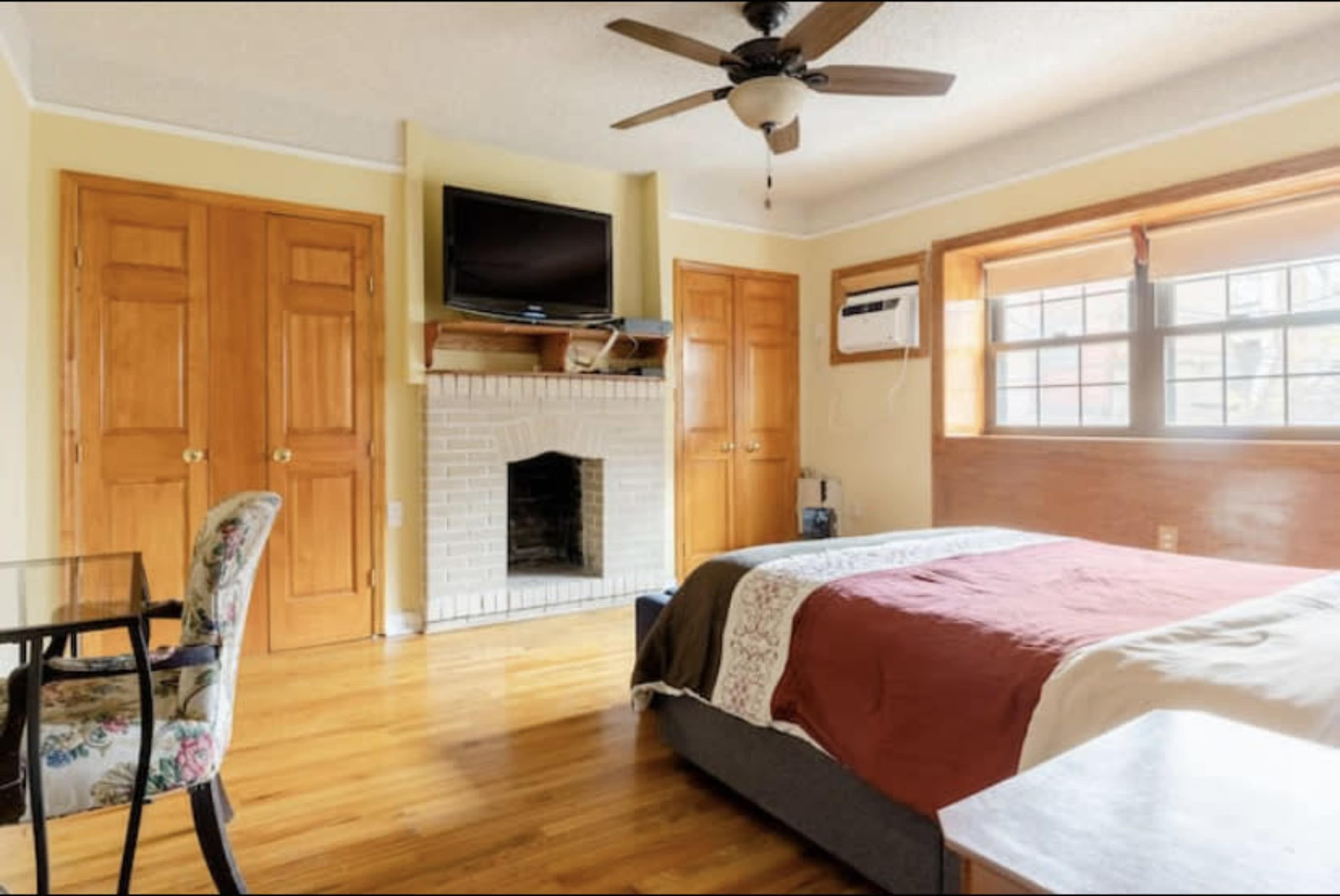 A bedroom features a bed with a red coverlet, a small dining table, a fireplace with a mounted TV above it, and double closets.