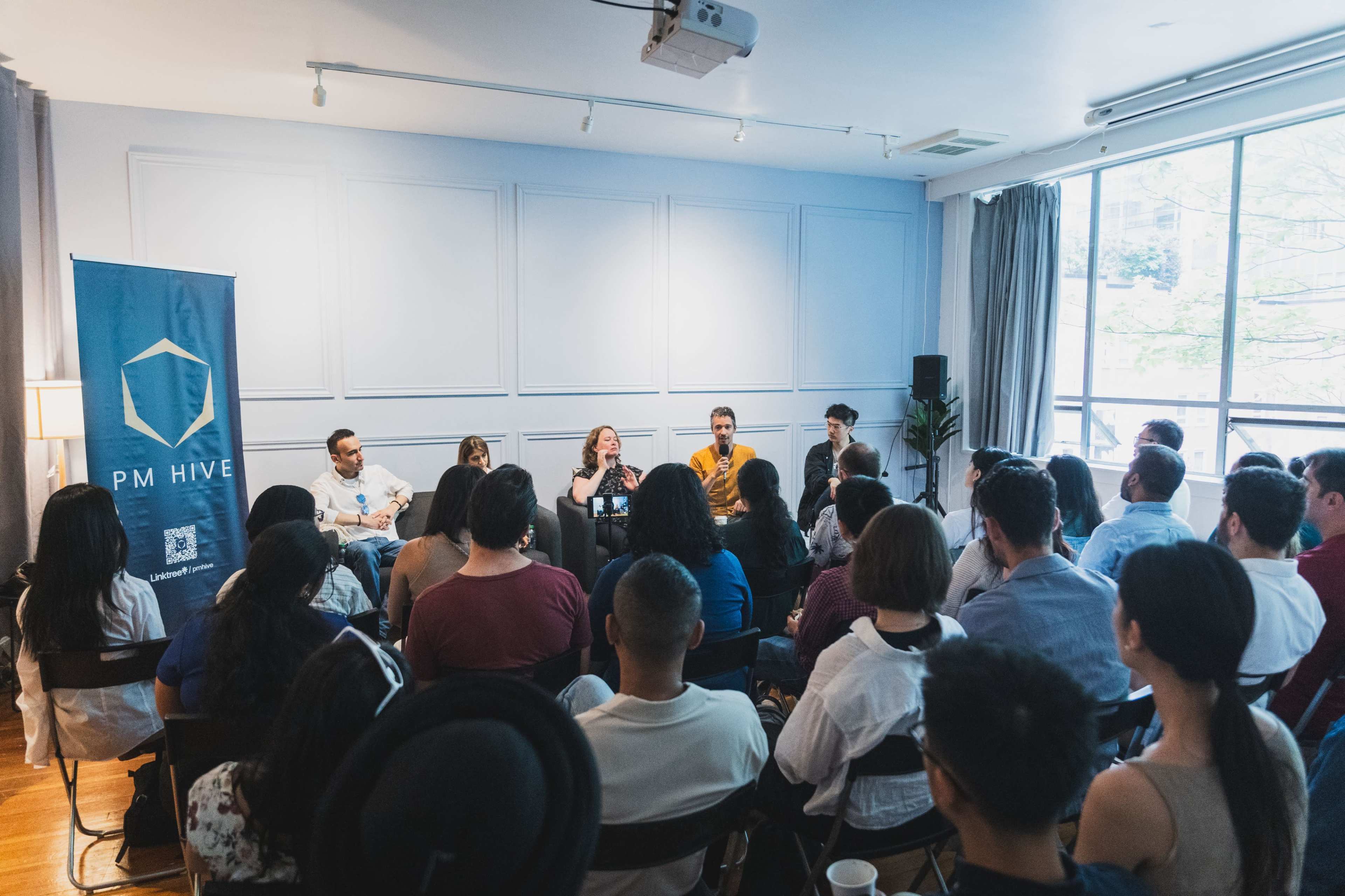 A panel discussion is taking place in a well-lit room, with an audience seated in front of a stage featuring a PM Hive banner.