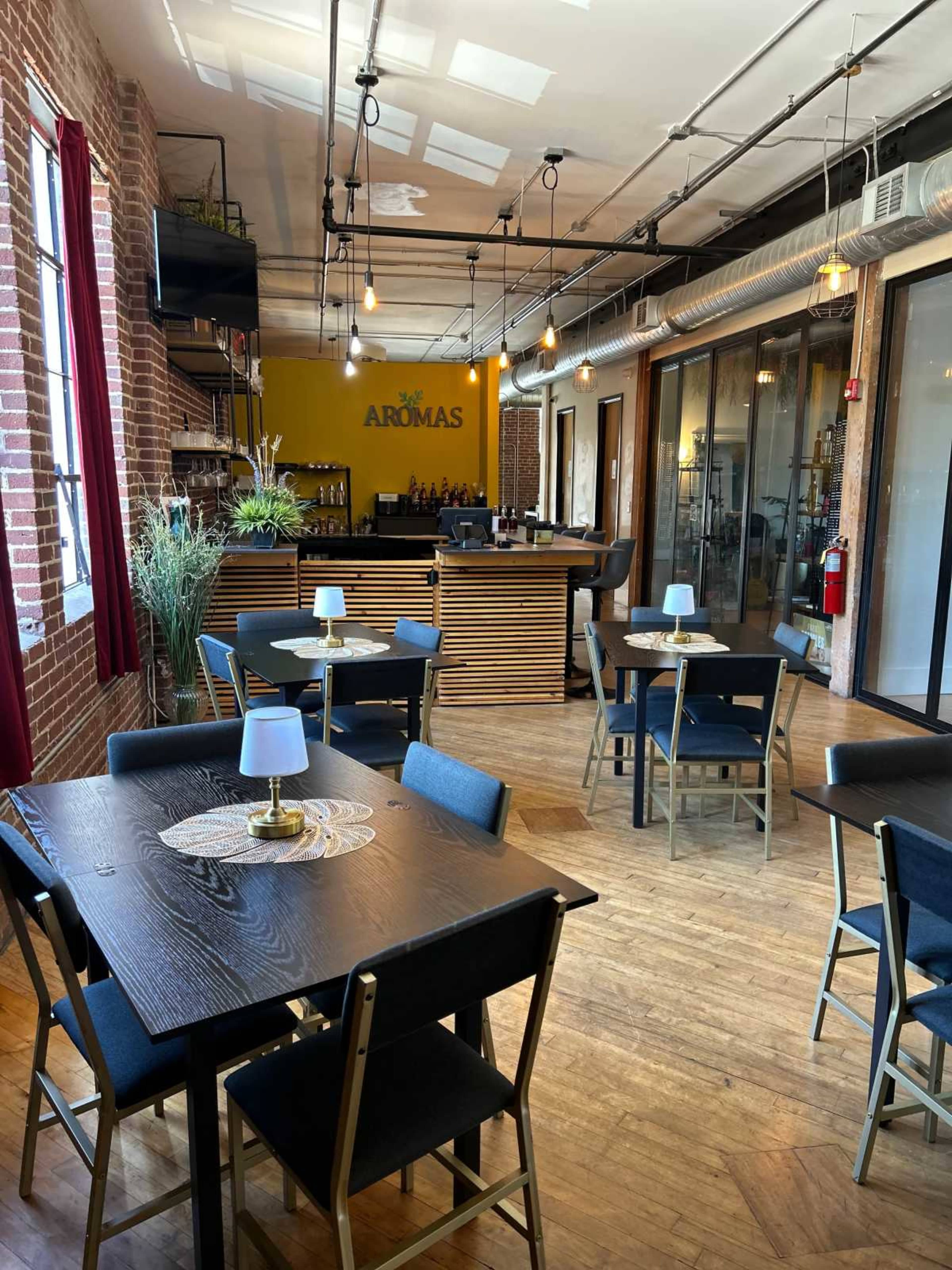 The image shows a modern dining area with wooden tables and chairs, illuminated by pendant lights, leading to a well-equipped bar in the background.