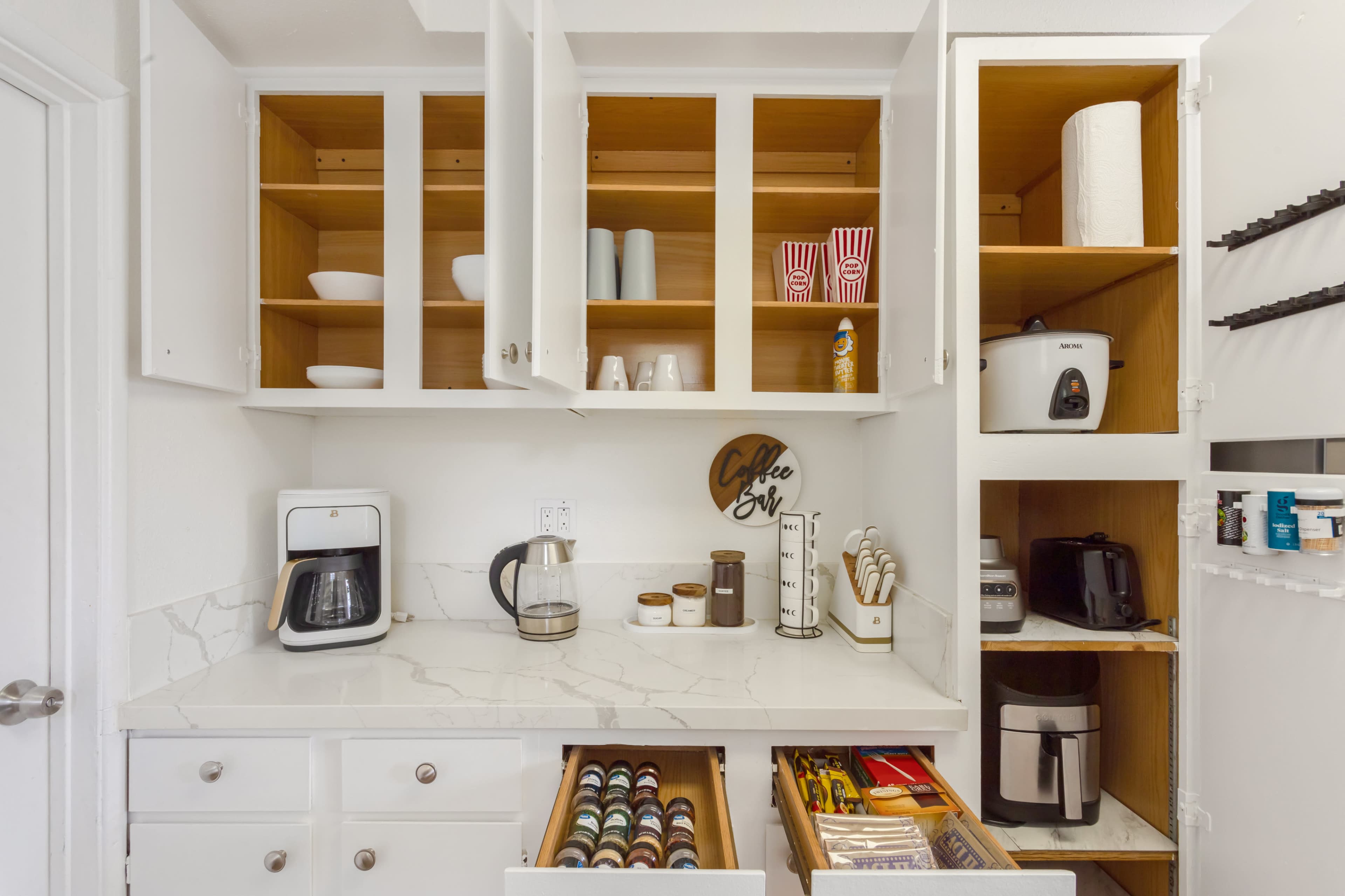 A kitchen cabinet area displays open shelves with dishes and snacks, a countertop coffee maker, and organized drawers containing various kitchen utensils and coffee pods.