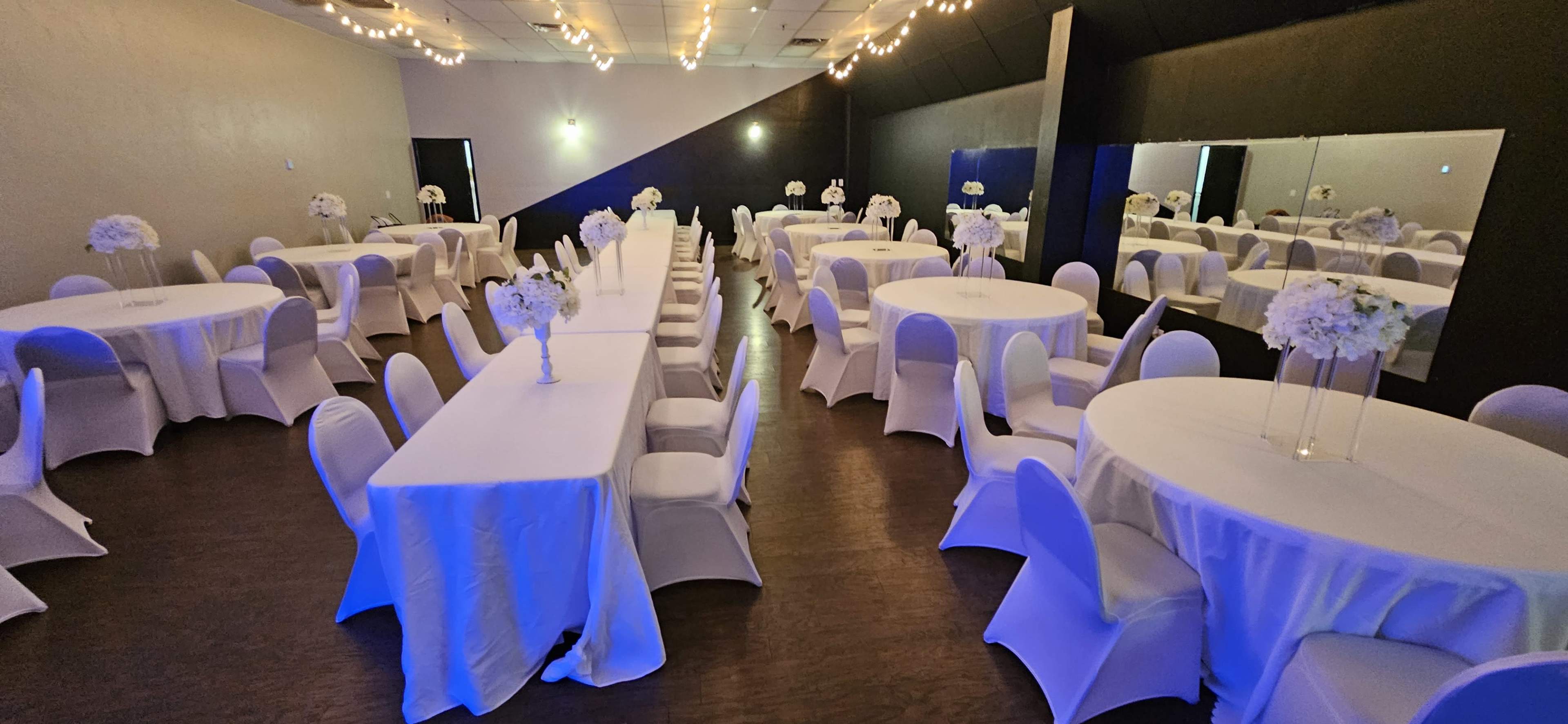 The image shows a banquet hall set up with several round tables covered in white tablecloths, decorated with floral centerpieces, and illuminated by soft lighting.
