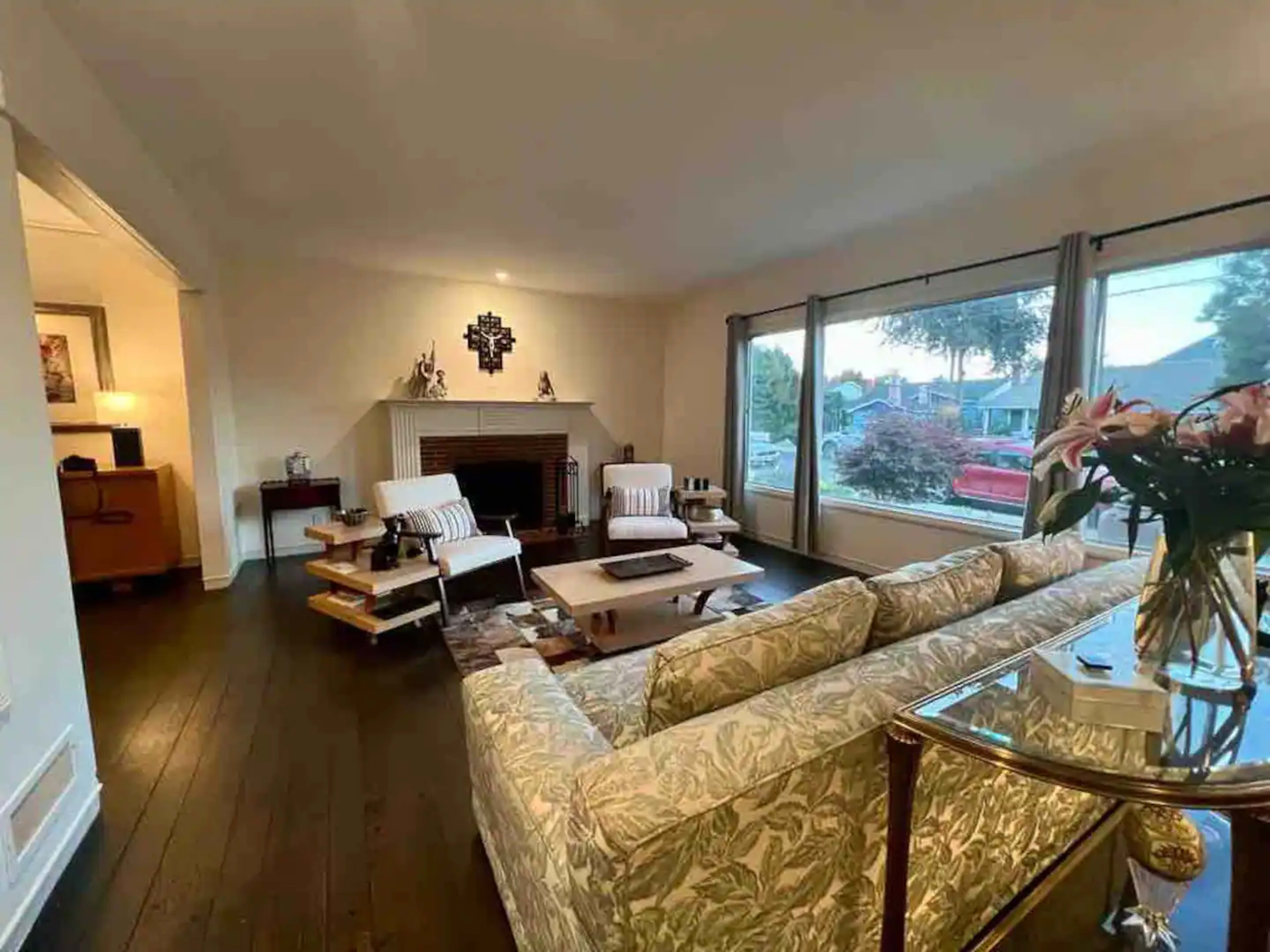 The living room features a floral-patterned sofa, two armchairs, a coffee table, and large windows overlooking a front yard.