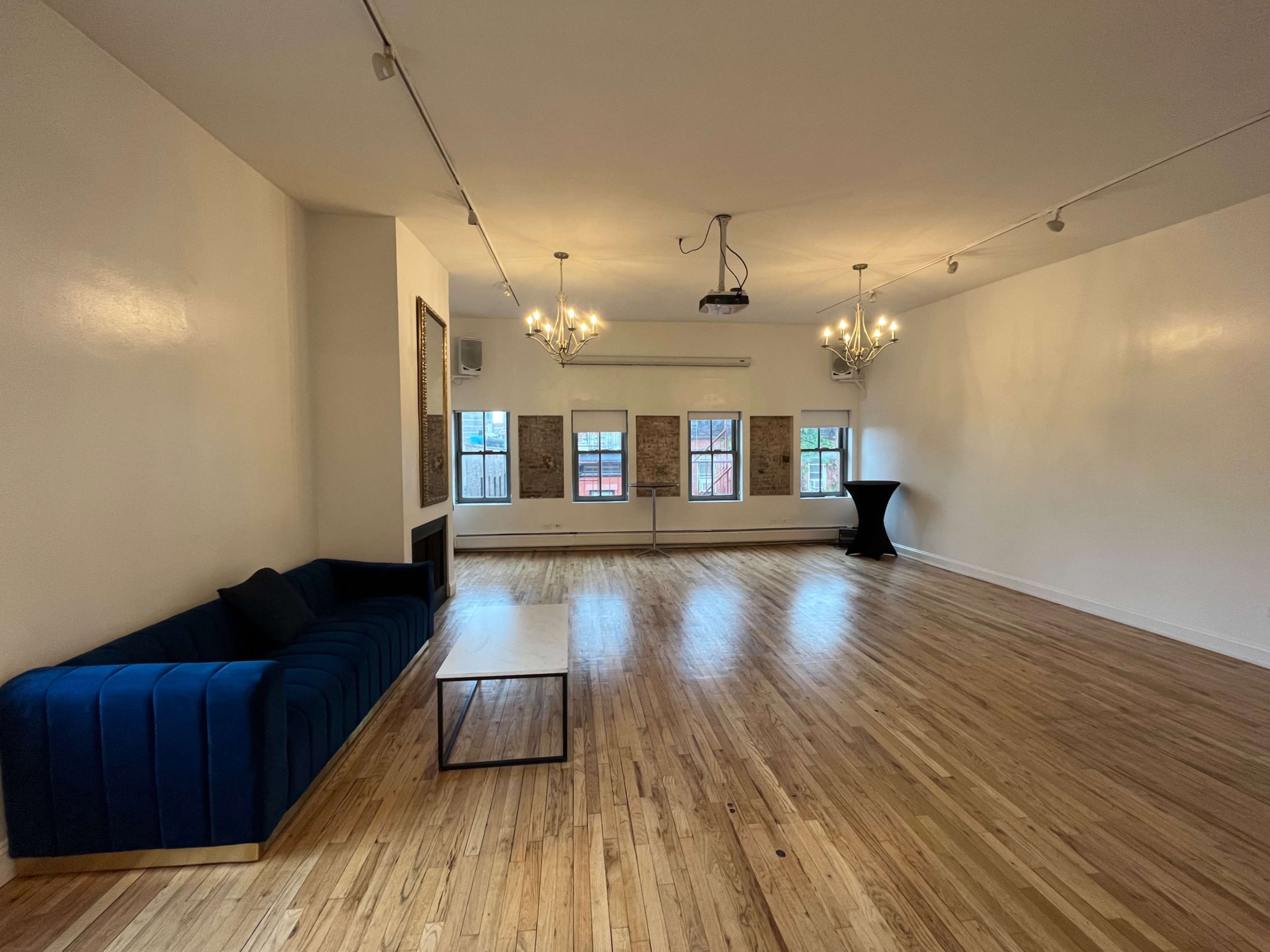 The image shows a spacious room with wooden floors, a blue sofa, a small coffee table, three windows, and two chandeliers.