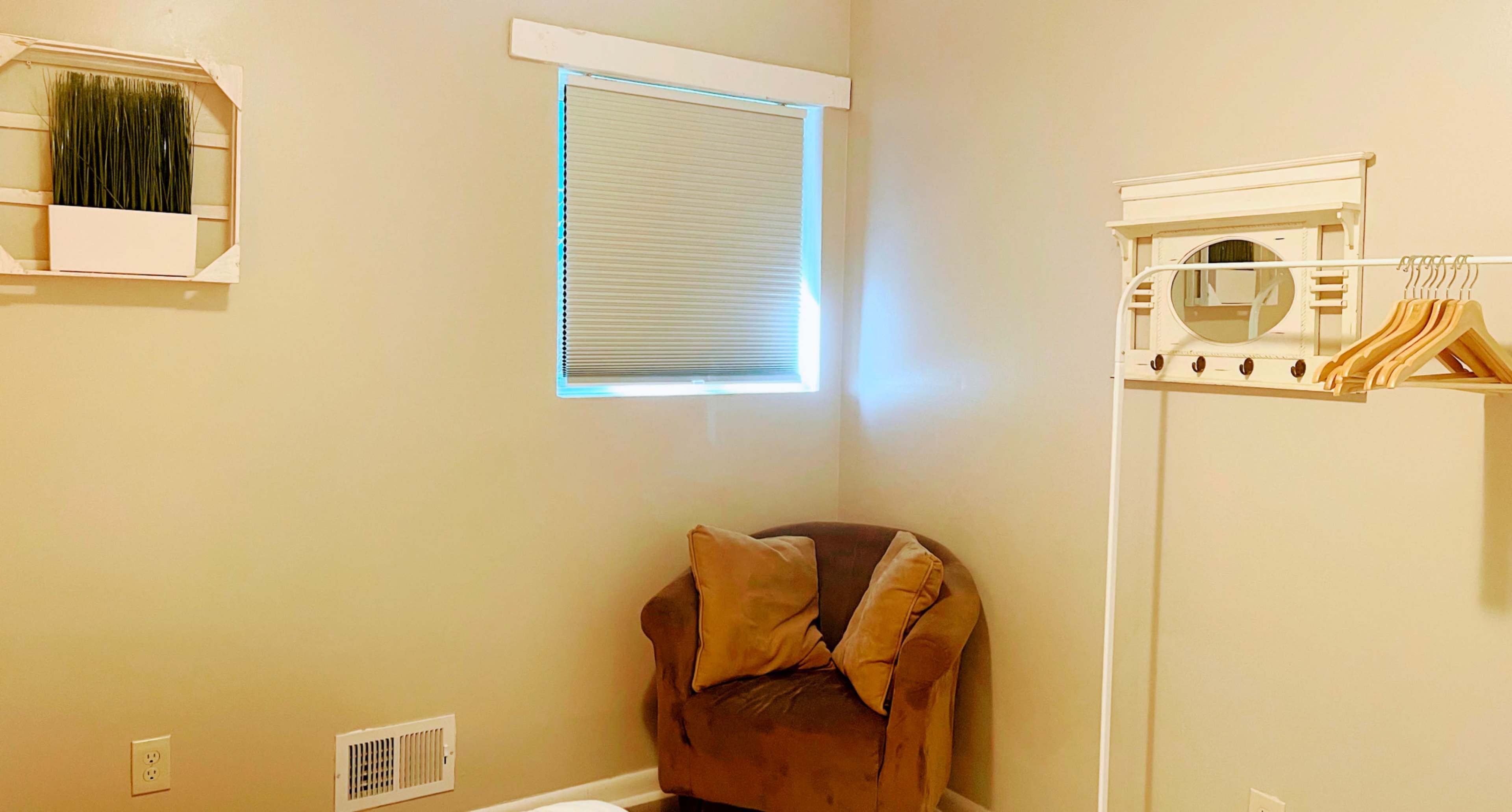 A small, light-colored room with a window, a brown chair in the corner, and a wall-mounted coat rack with wooden hangers.