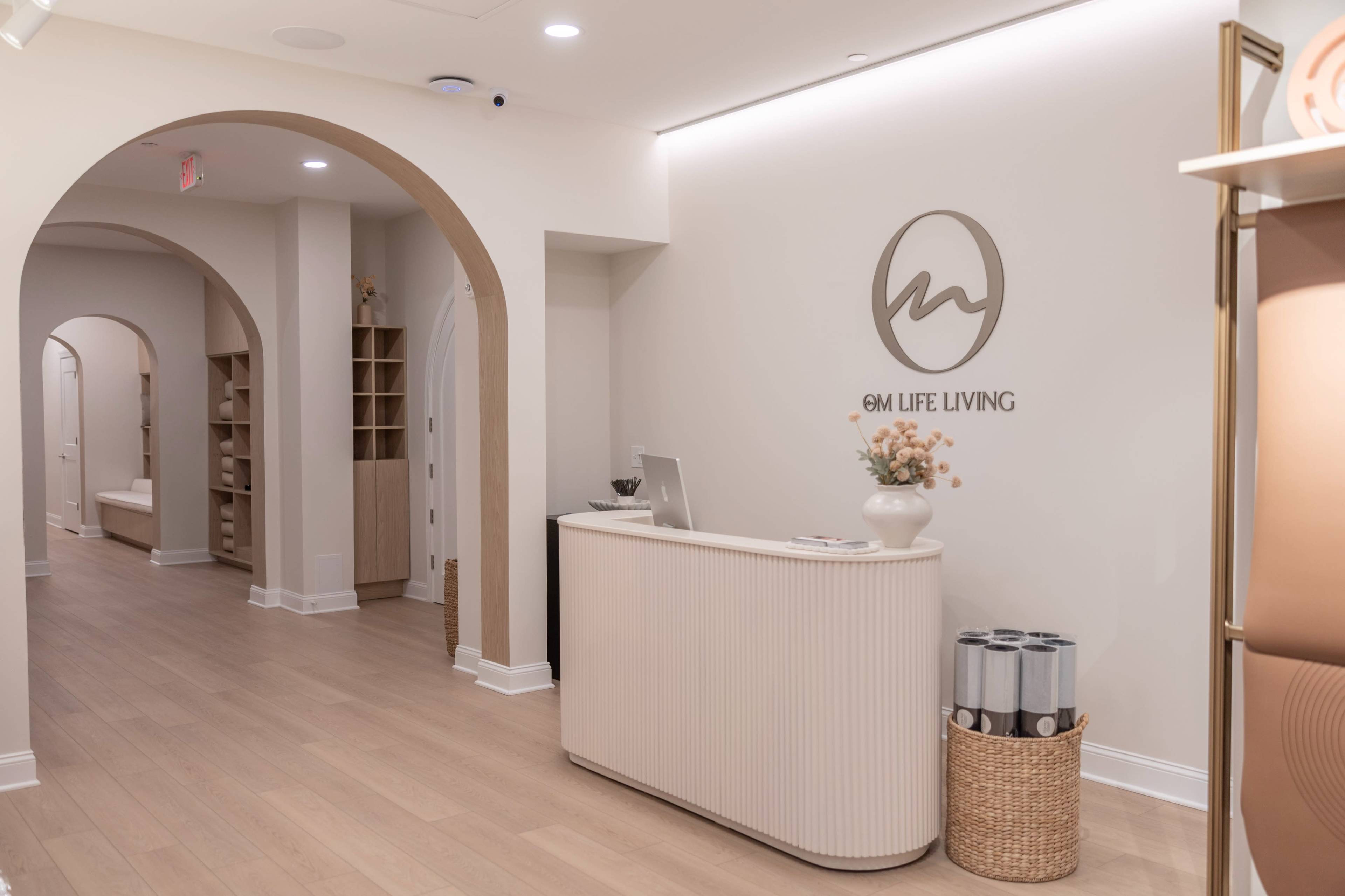 The image shows a modern reception area with a curved desk, a logo on the wall, and arched doorways leading to a hallway in a well-lit interior space.