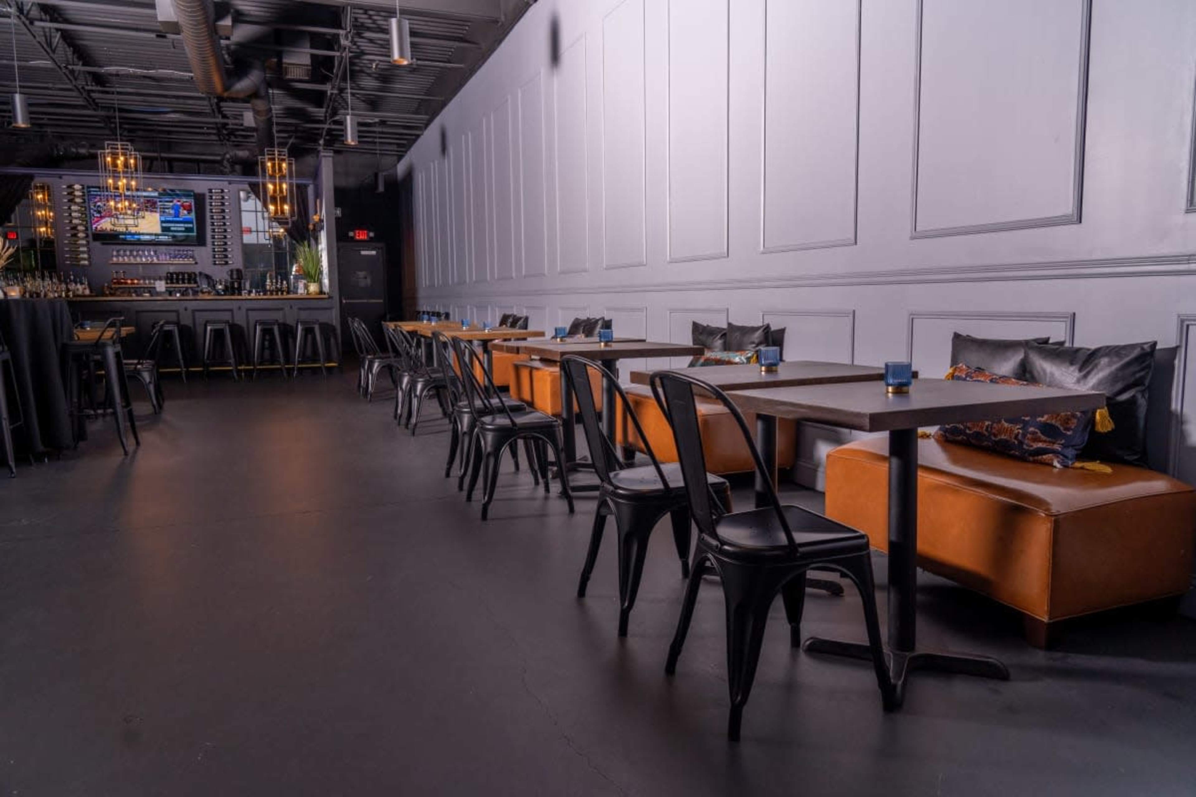 A modern interior of a restaurant featuring rows of black chairs and tables along a wall, with a bar area illuminated in the background.