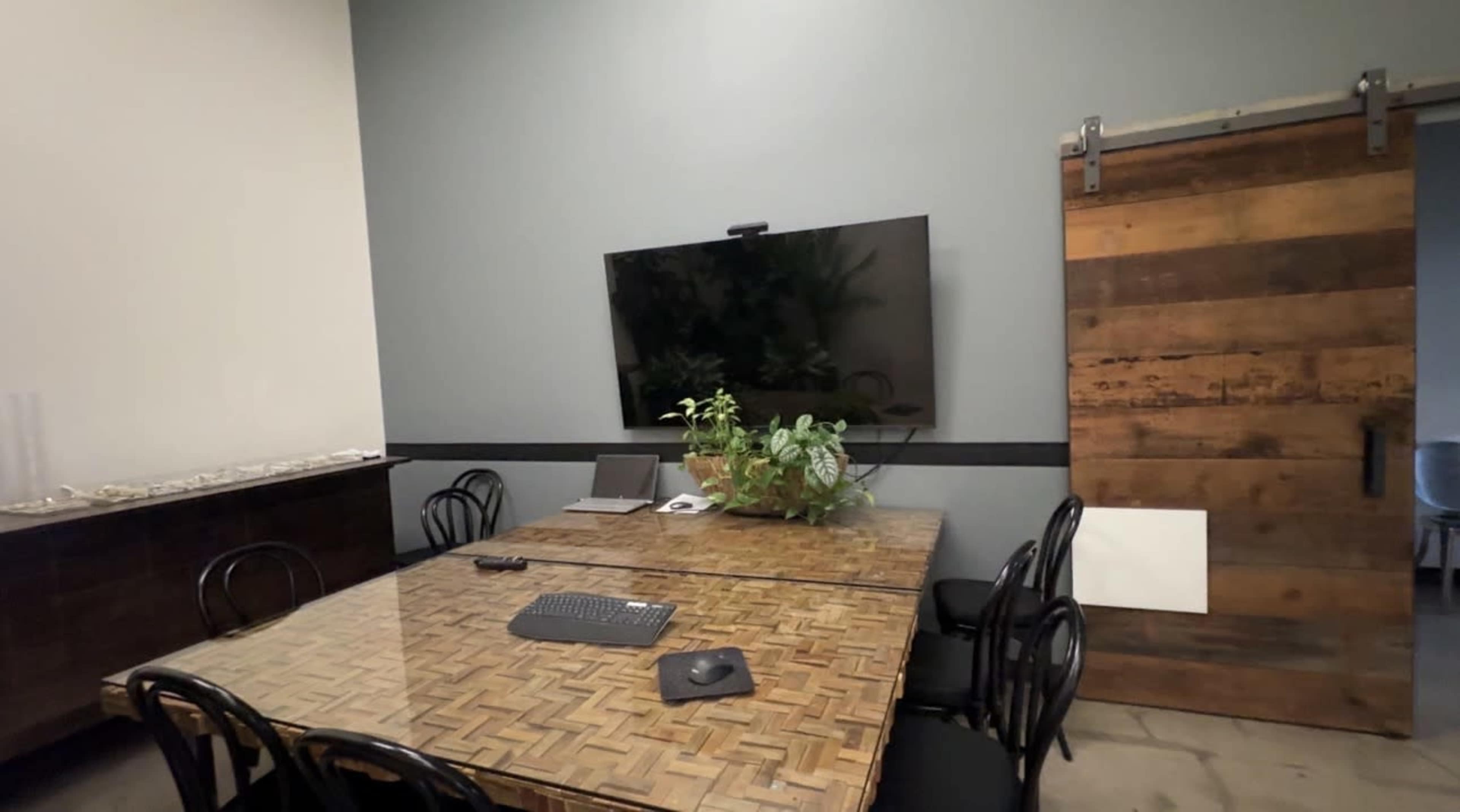 A conference room features a large wooden table, black chairs, a television mounted on the wall, and a sliding barn door.