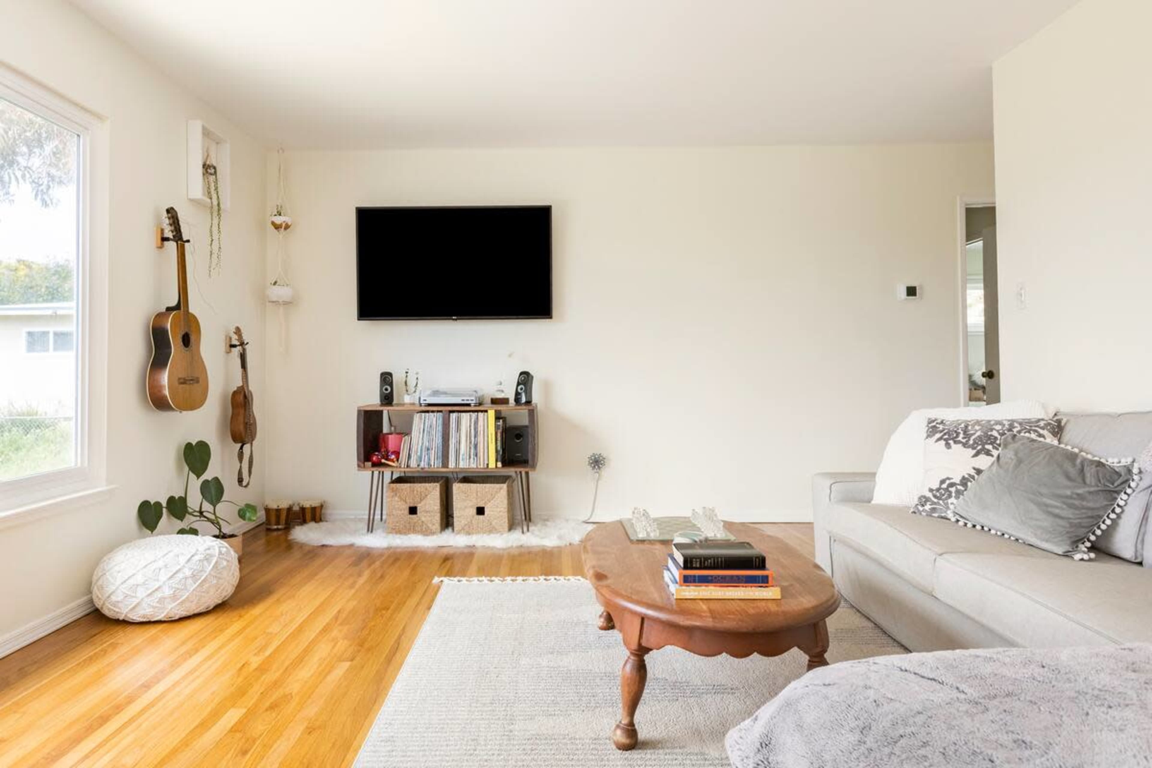 A bright living room featuring a light-colored sofa, a wooden coffee table, a television mounted on the wall, and a shelving unit filled with books and storage baskets.