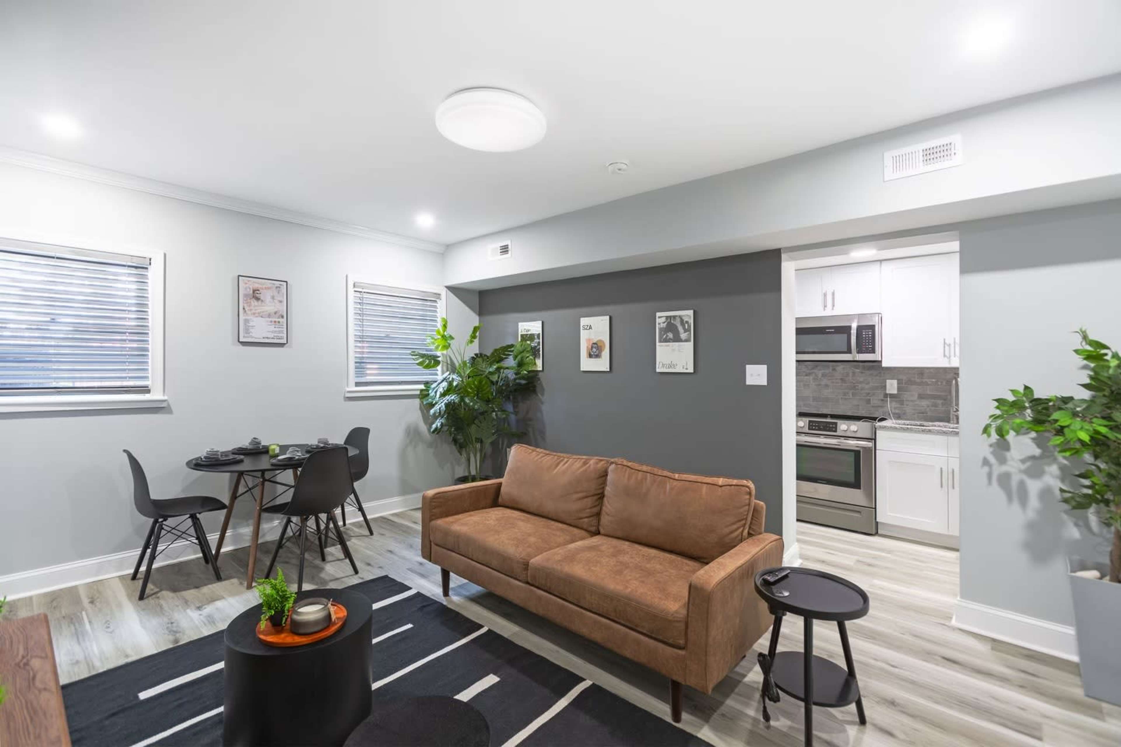 A modern living area with a brown sofa, a small round table with chairs, and a kitchen area visible in the background.