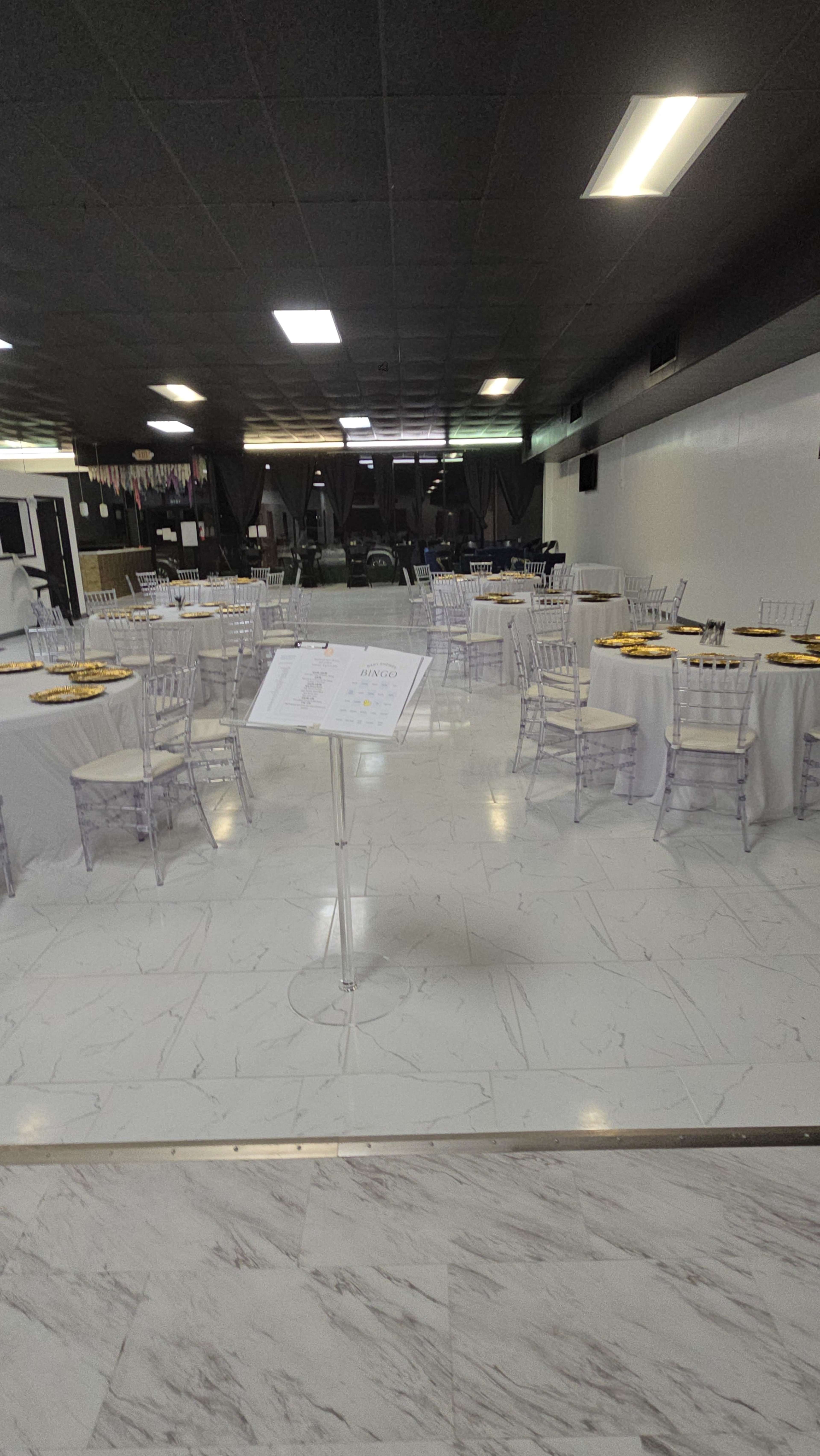 A banquet hall with several round tables set with white tablecloths and clear chairs, along with a sign in the foreground and circular trays of food on the tables.