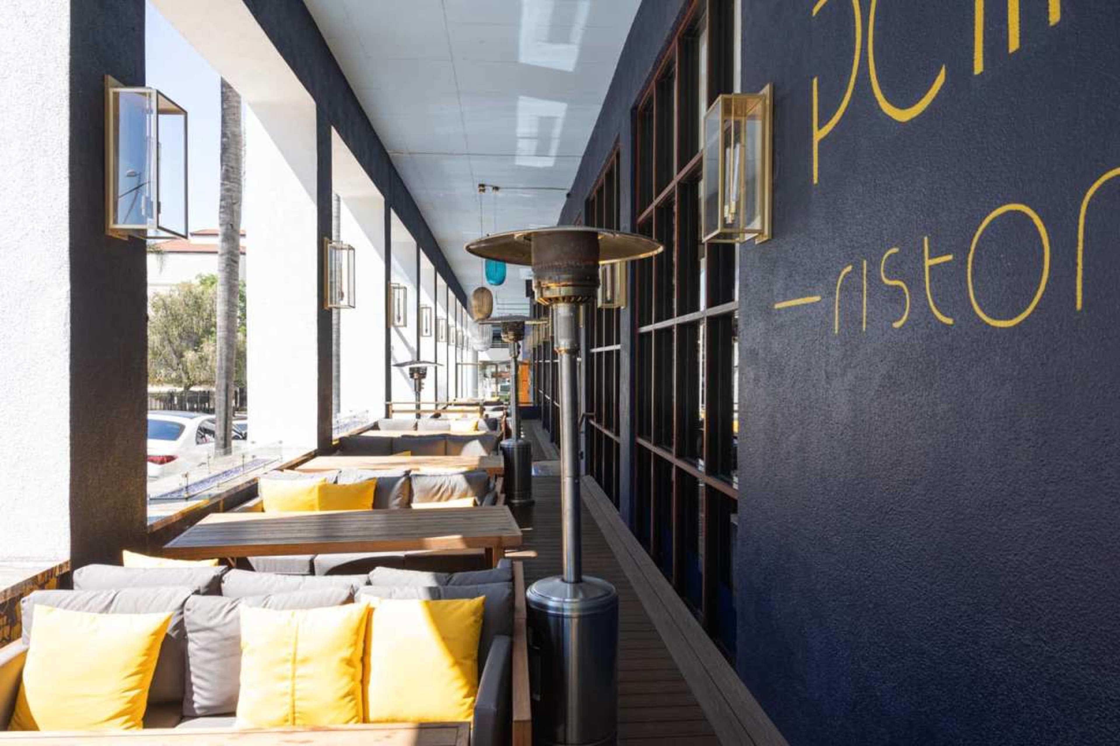 The image shows a restaurant patio featuring wooden seating with yellow cushions, outdoor heaters, and large windows.