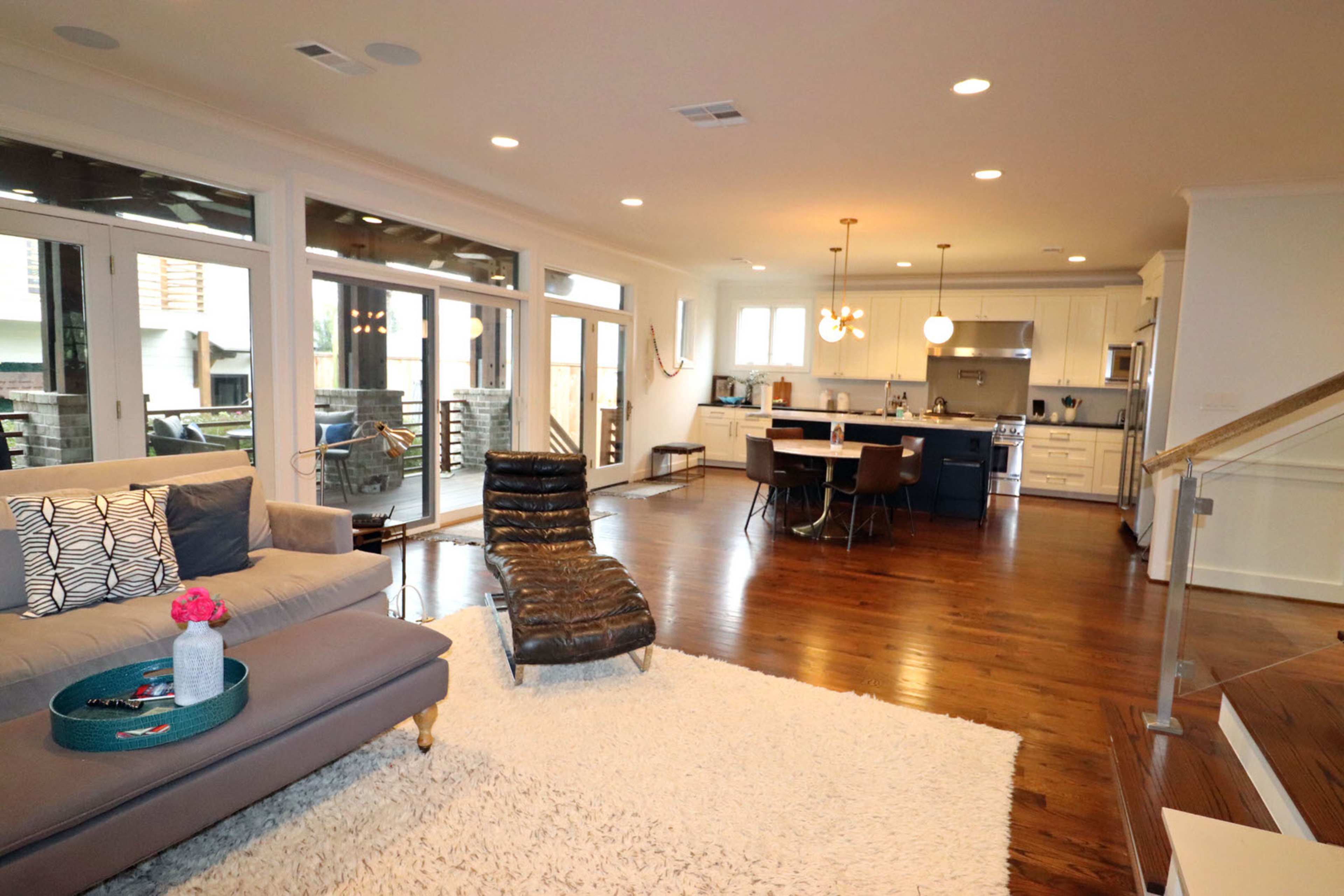 A modern living room and kitchen area featuring hardwood floors and large windows that illuminate the space.