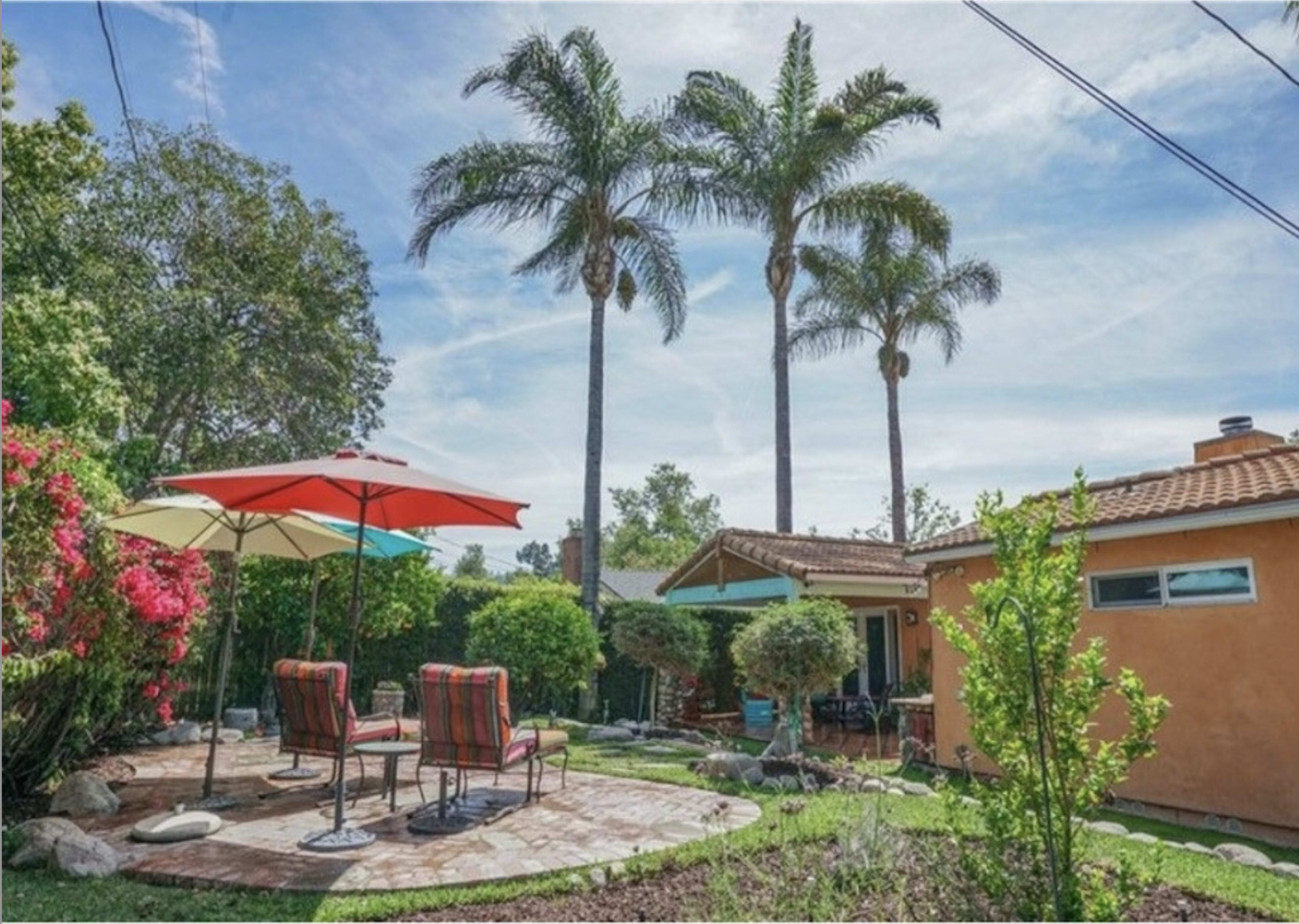 The image shows a landscaped backyard with a stone patio, two chairs, colorful umbrellas, and palm trees in the background.