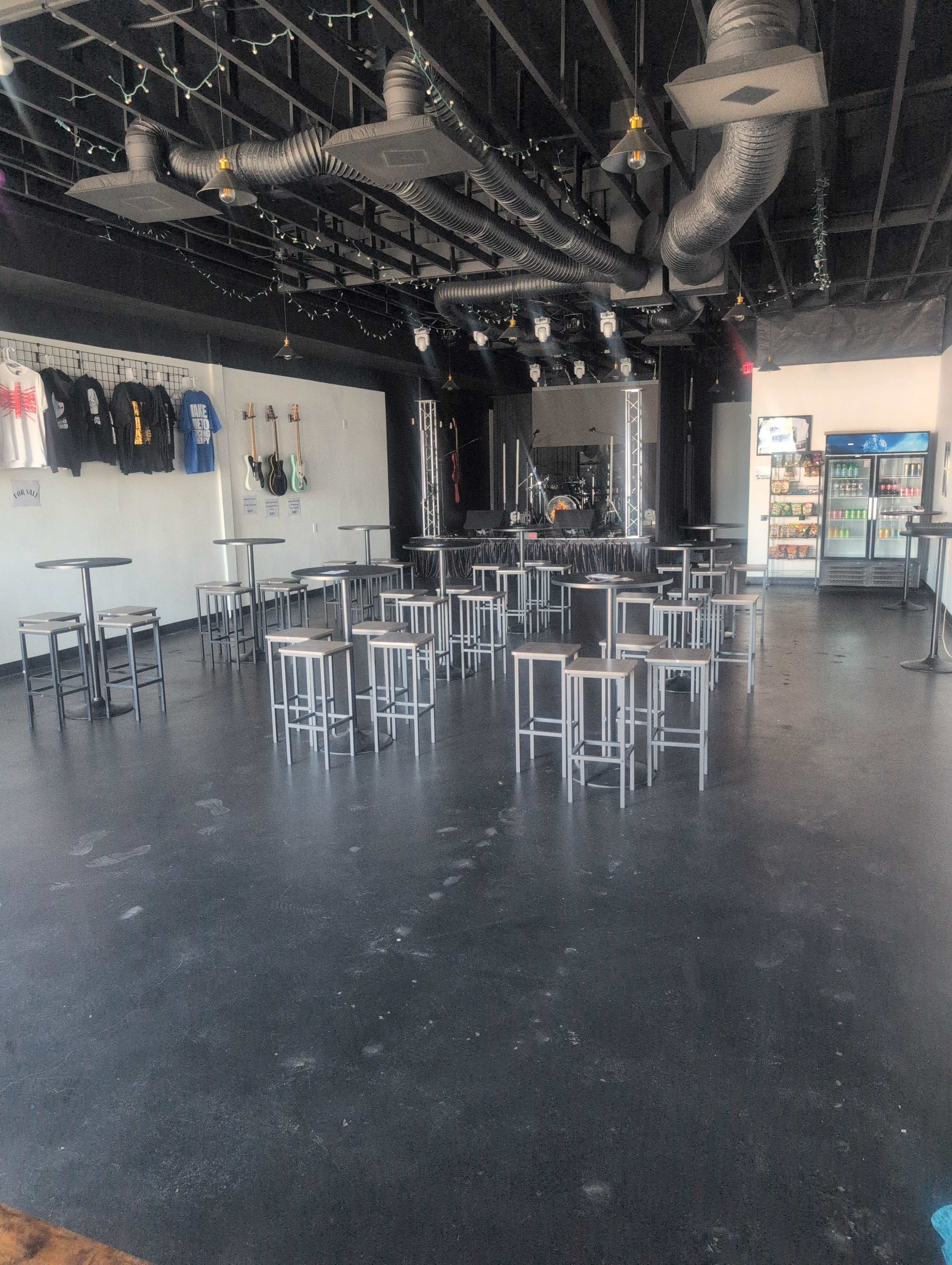 The image shows an empty music venue with rows of metal stools arranged around small tables, and a stage with musical instruments in the background.