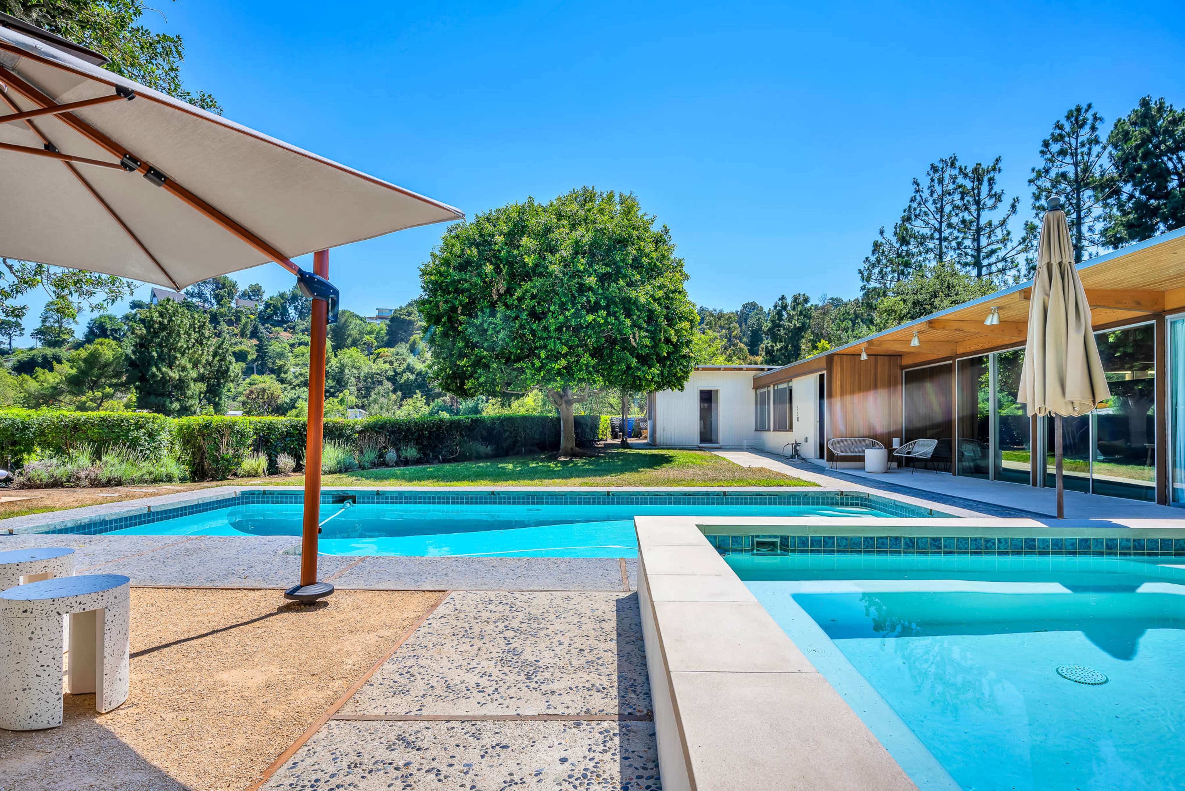 A modern backyard features a swimming pool, a shaded seating area under an umbrella, and a contemporary home with large windows.