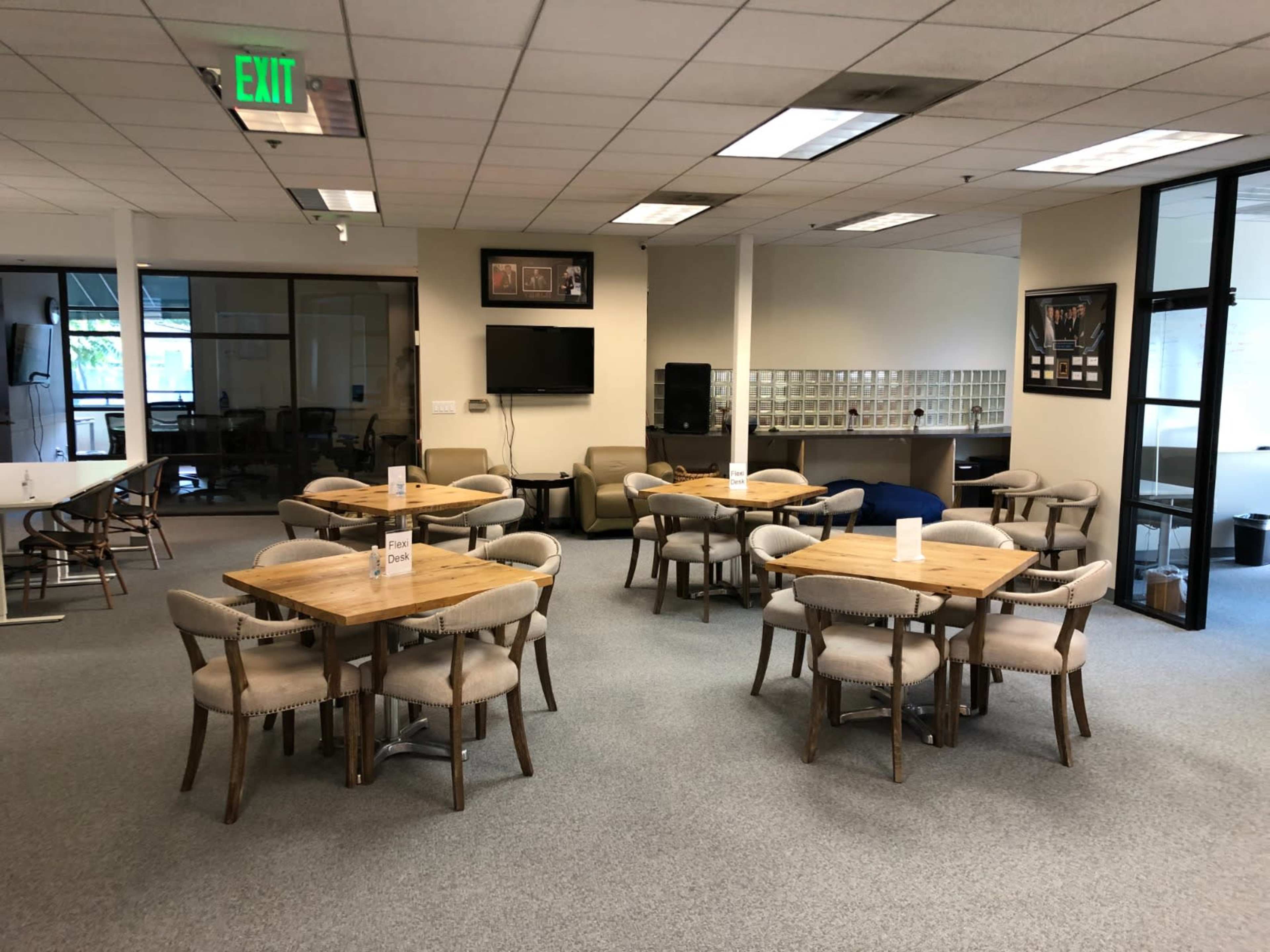 A spacious room features several wooden tables surrounded by chairs, with a television and seating area in the background.