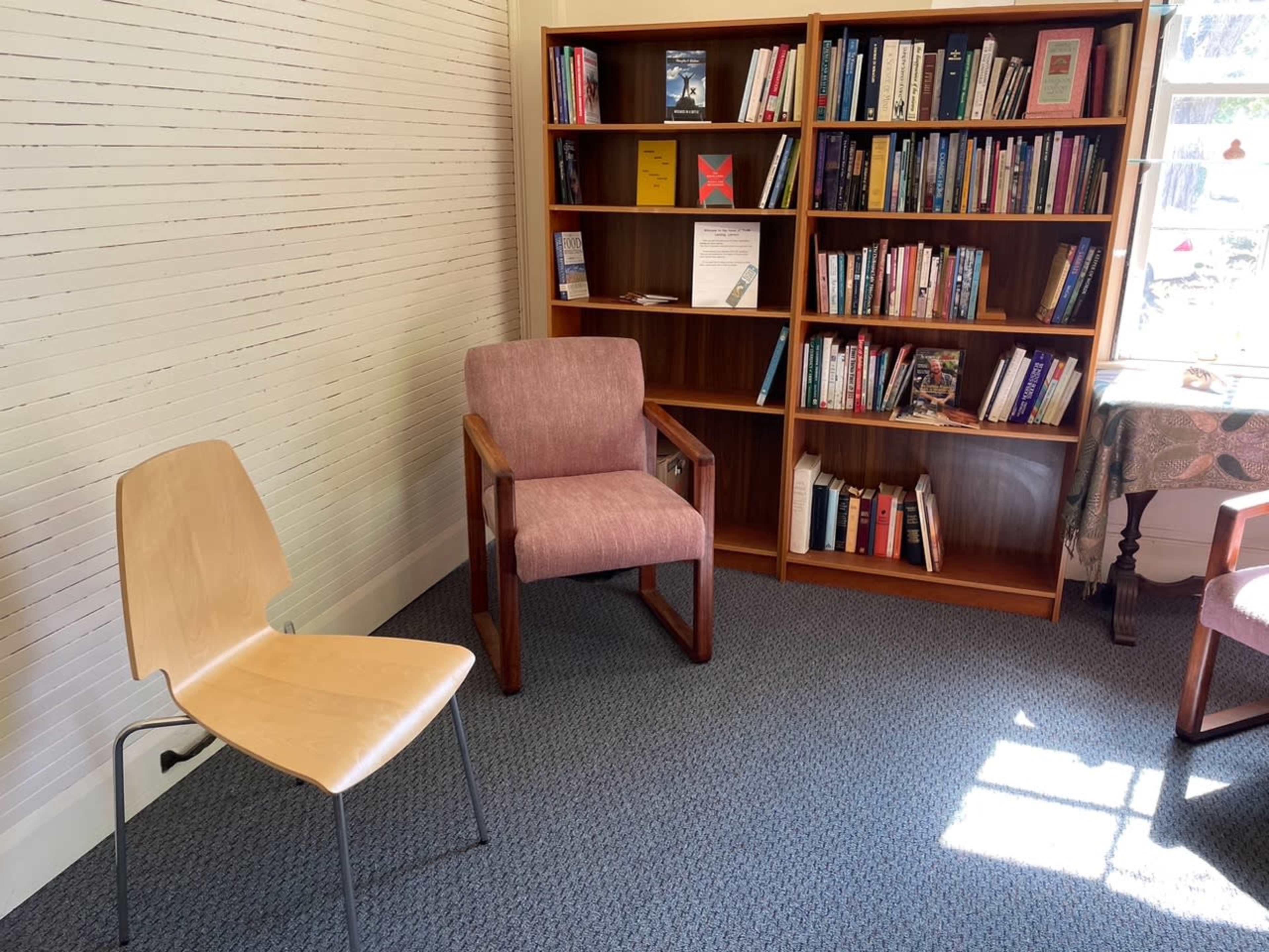 The image shows a room with two chairs, a wooden bookshelf filled with books, and natural light coming through a window.