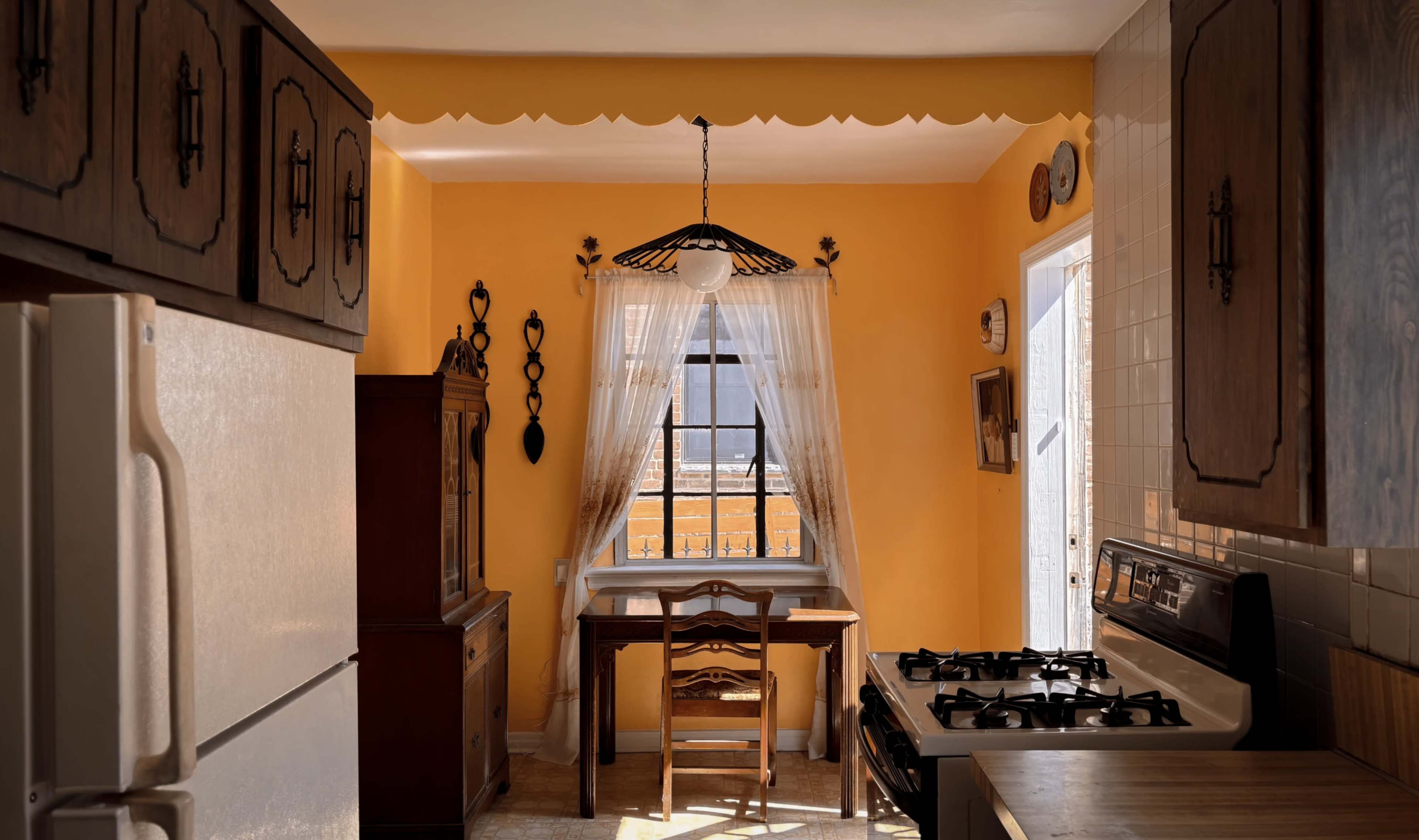 The image depicts a warm-toned kitchen featuring wooden cabinets, a dining table beside a window, and a stove against a wall.