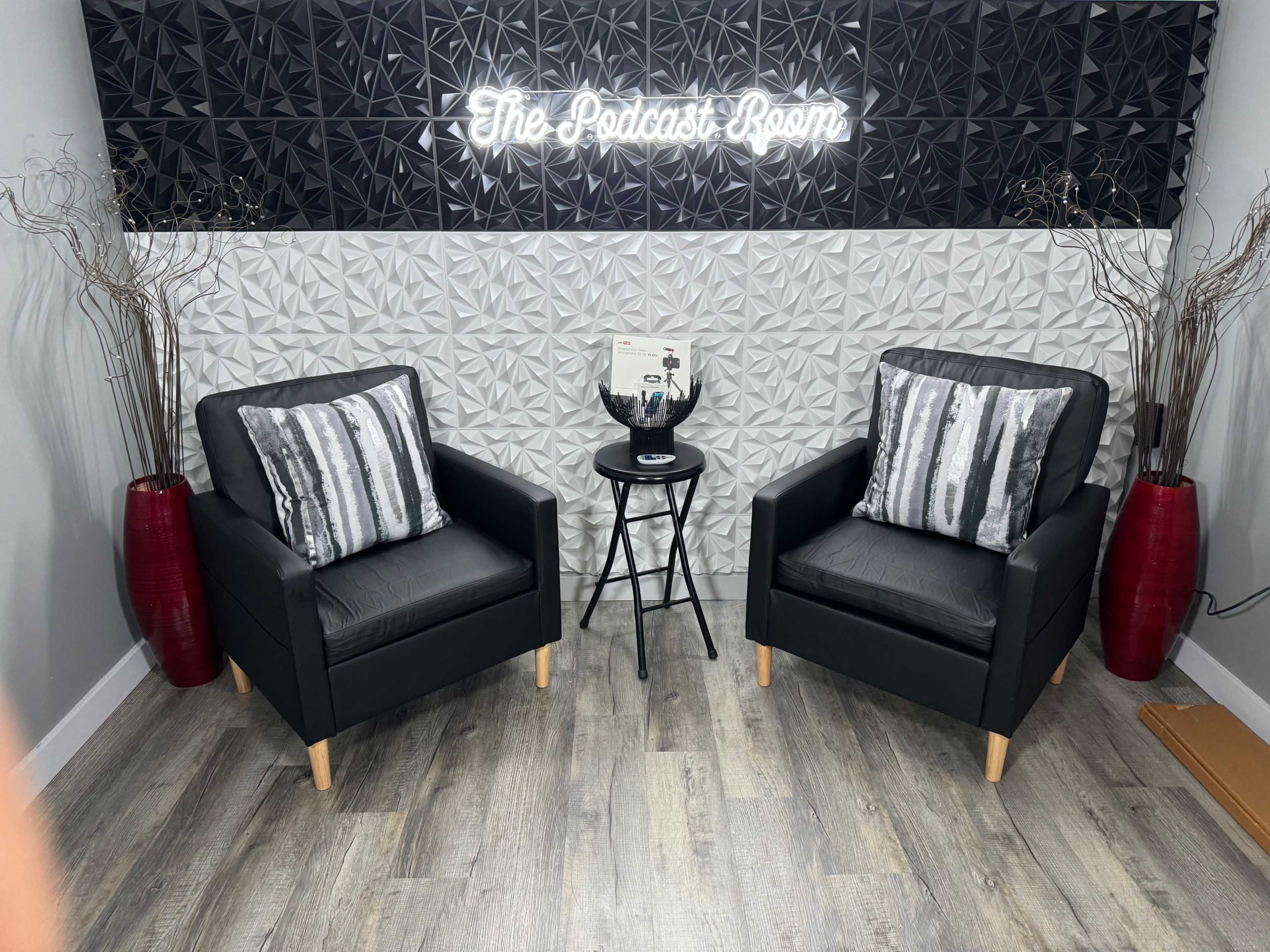 The image shows two black armchairs with striped cushions and a small black table between them, set against a textured wall featuring the text "The Podcast Room" in neon lighting.