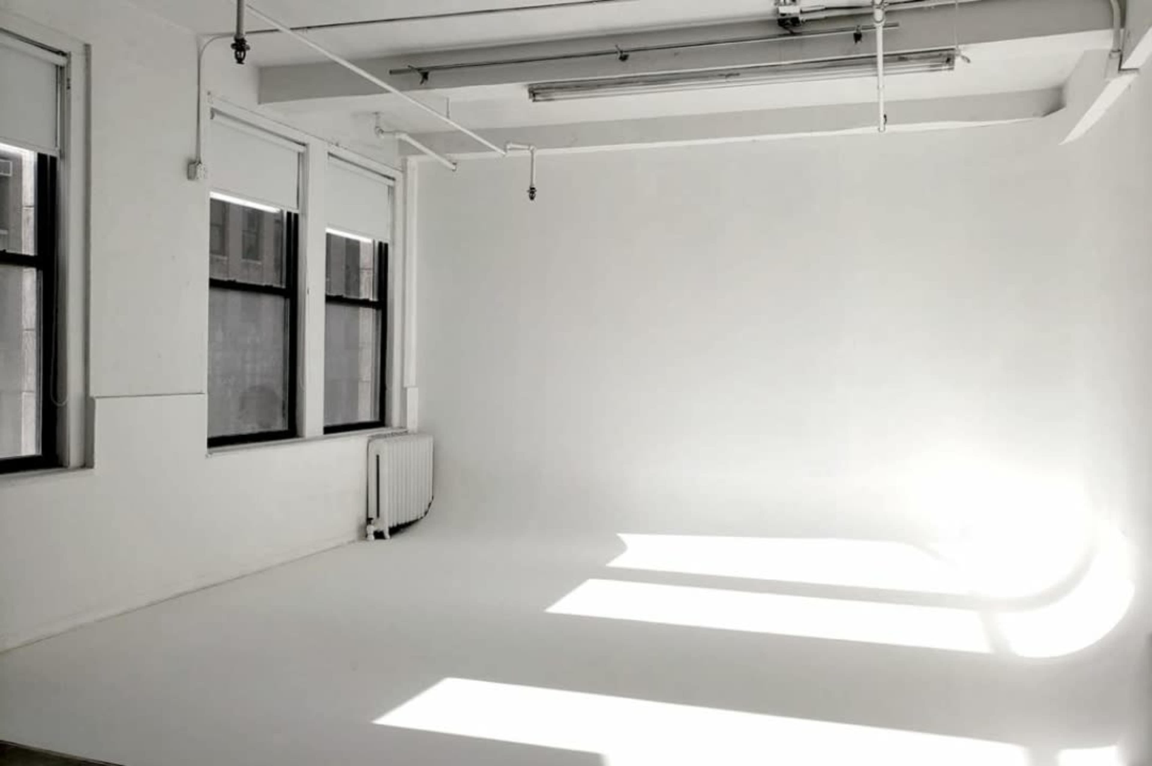 A bright, empty room with white walls and a seamless white floor, illuminated by sunlight streaming through large windows.