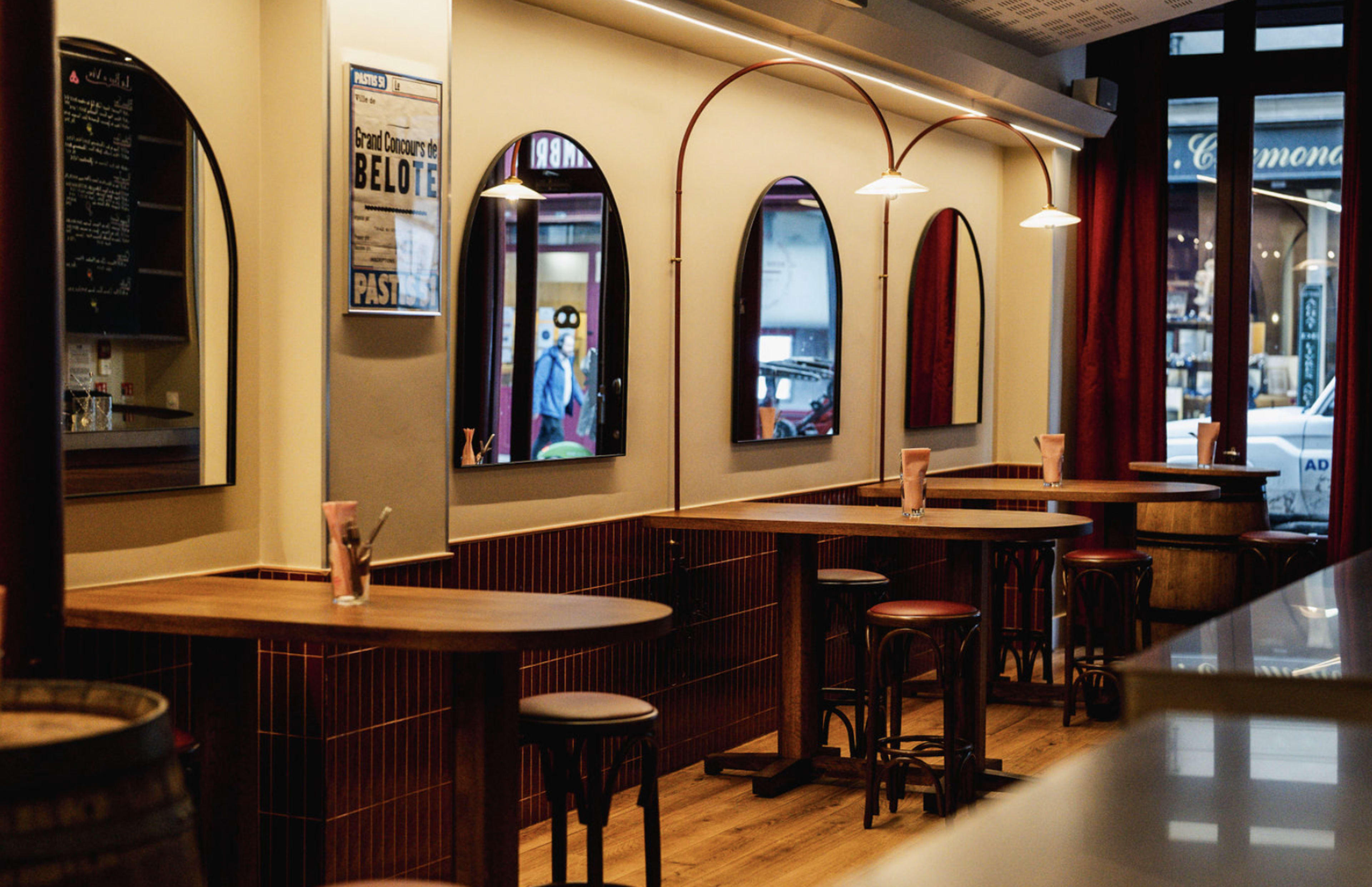 The image shows a modern bar area with wooden tables and stools, mirrored arches on the walls, and a soft-lit interior.