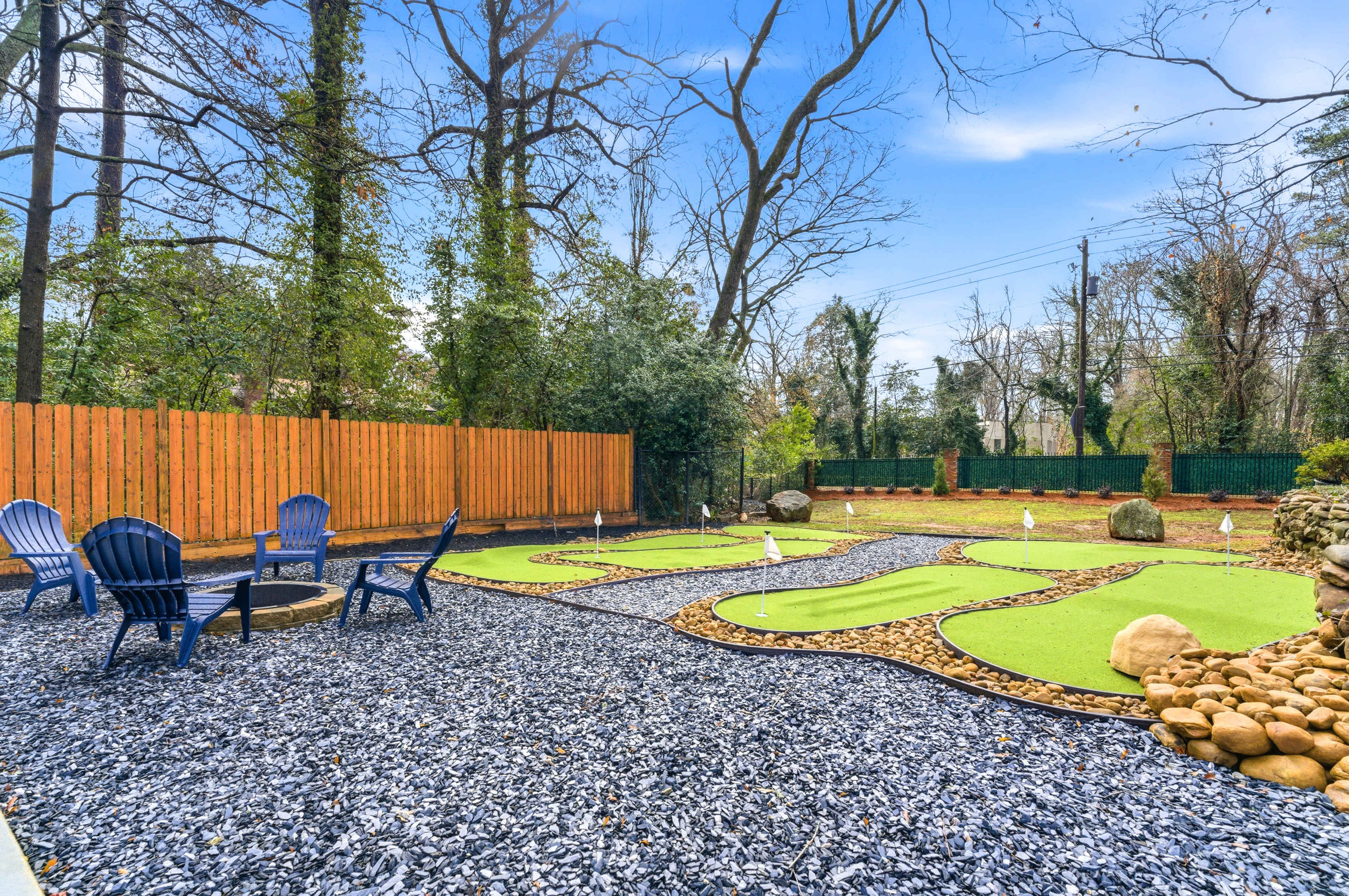 The image shows a mini-golf area with two putting greens surrounded by gravel, seating chairs, and a wooden fence in a wooded setting.