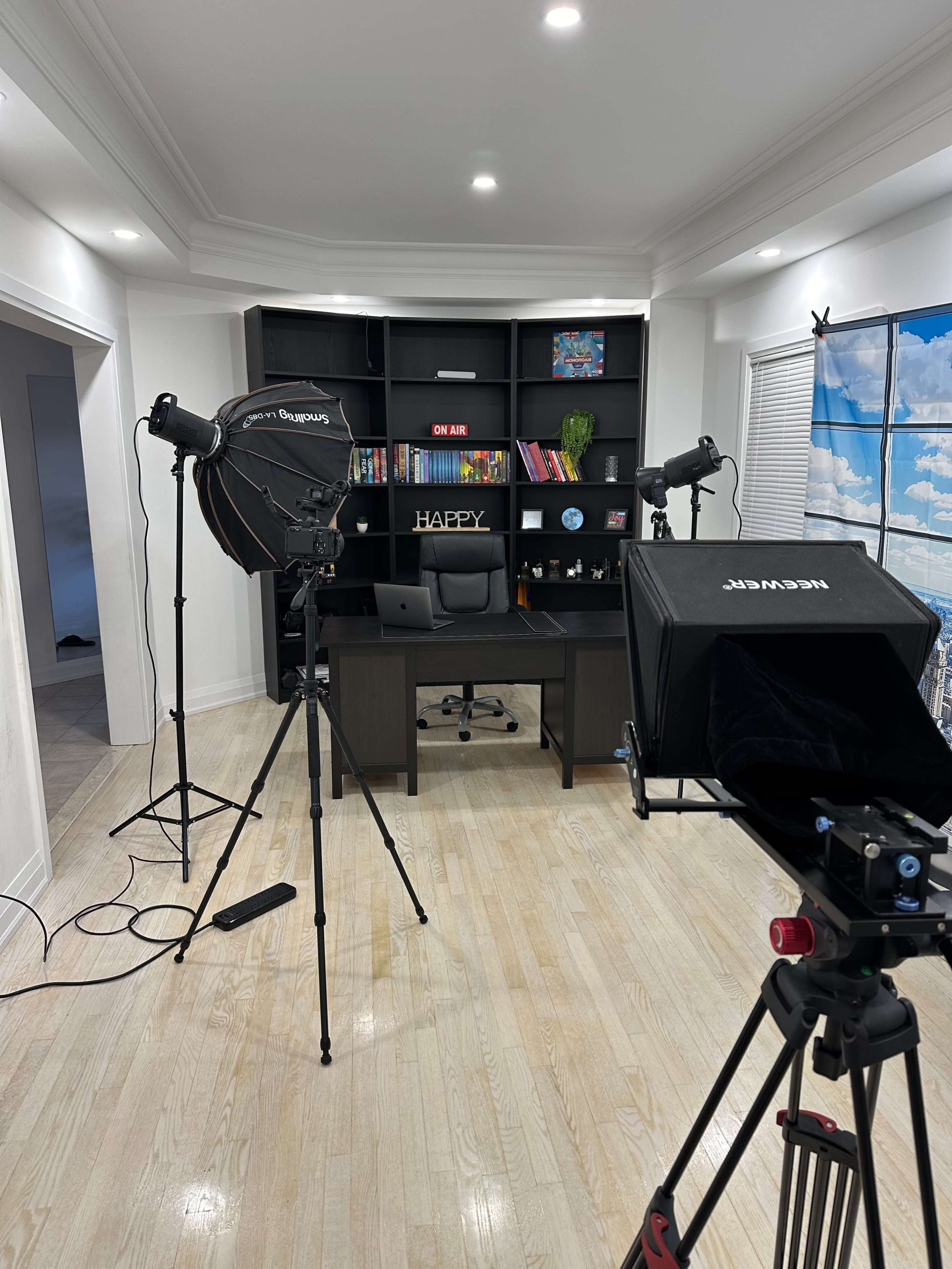 The image shows a well-lit room with a desk and bookshelves, equipped with lighting setups and camera equipment for a video production.